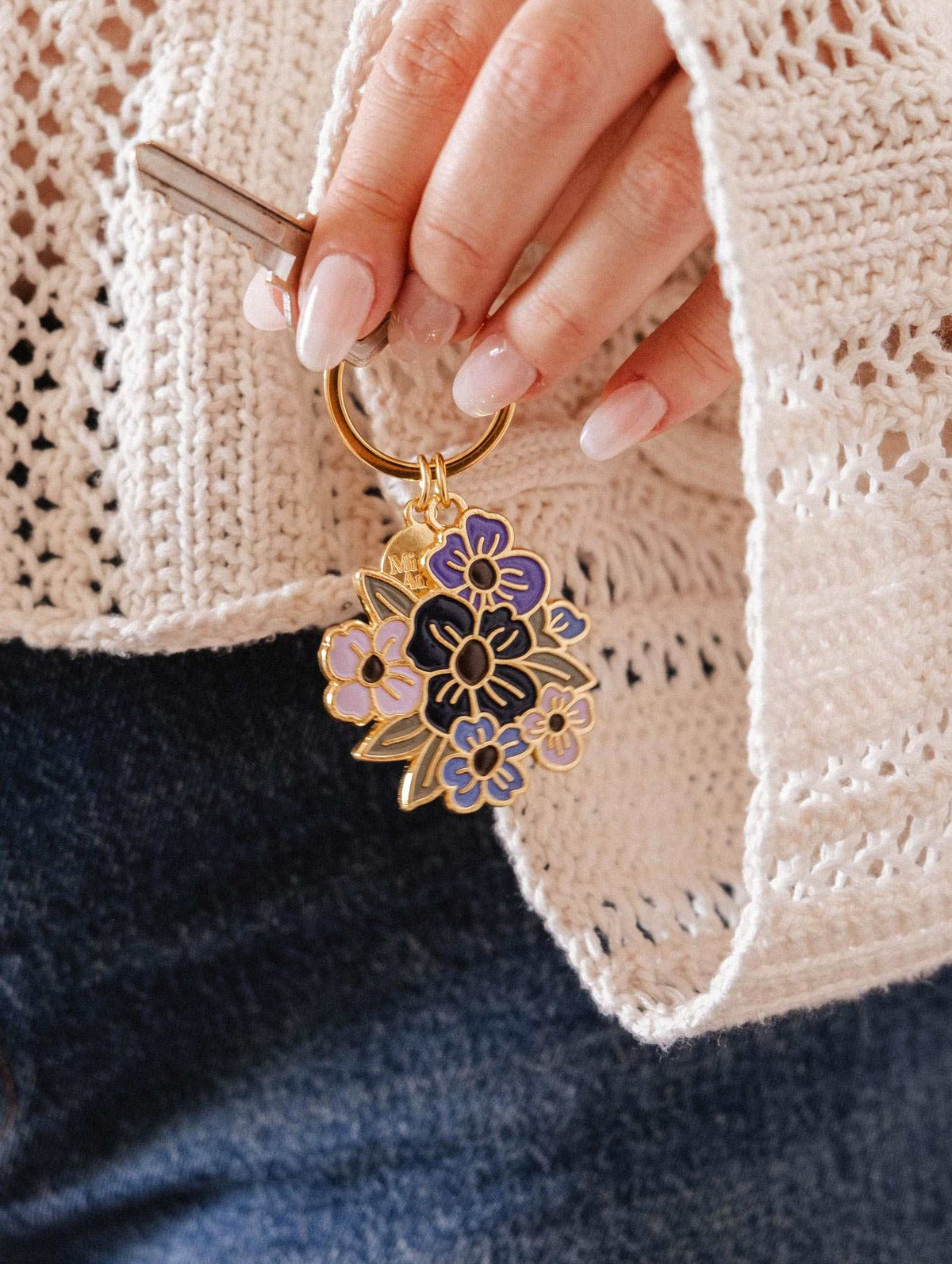 A hand holds the Mimi & August Floral Bouquet Keychain, showcasing purple and black enamel flowers, paired with a cream knit sweater and blue jeans. This Montreal-designed accessory adds a charming touch to any outfit.