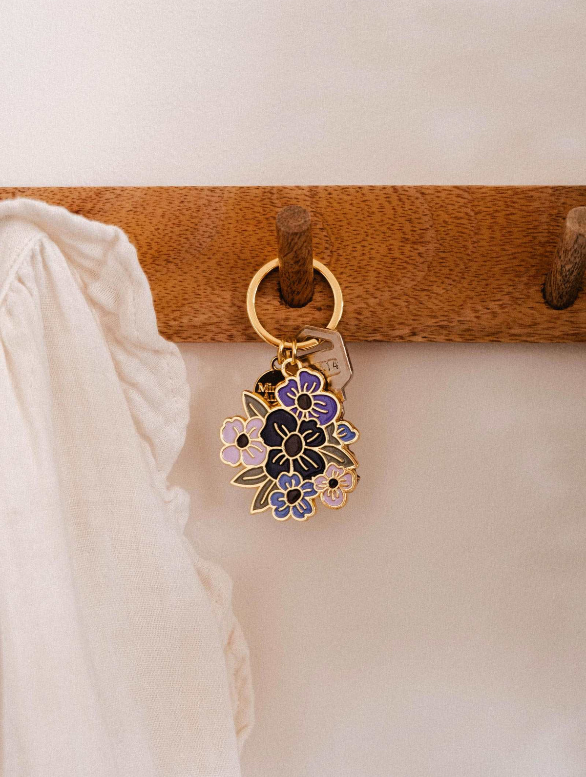 A gold Floral Bouquet Keychain from Mimi & August, featuring a purple and black enamel design, hangs on a wooden hook beside a ruffled white fabric—a charming accessory designed in Montreal.