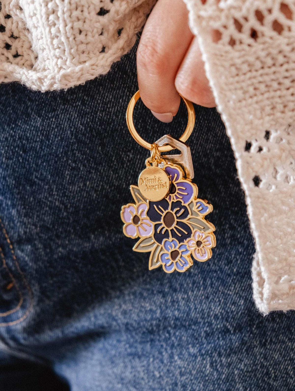 A hand holds Mimi & August's Floral Bouquet Keychain and two keys, set against blue denim jeans and a cream crochet sweater.