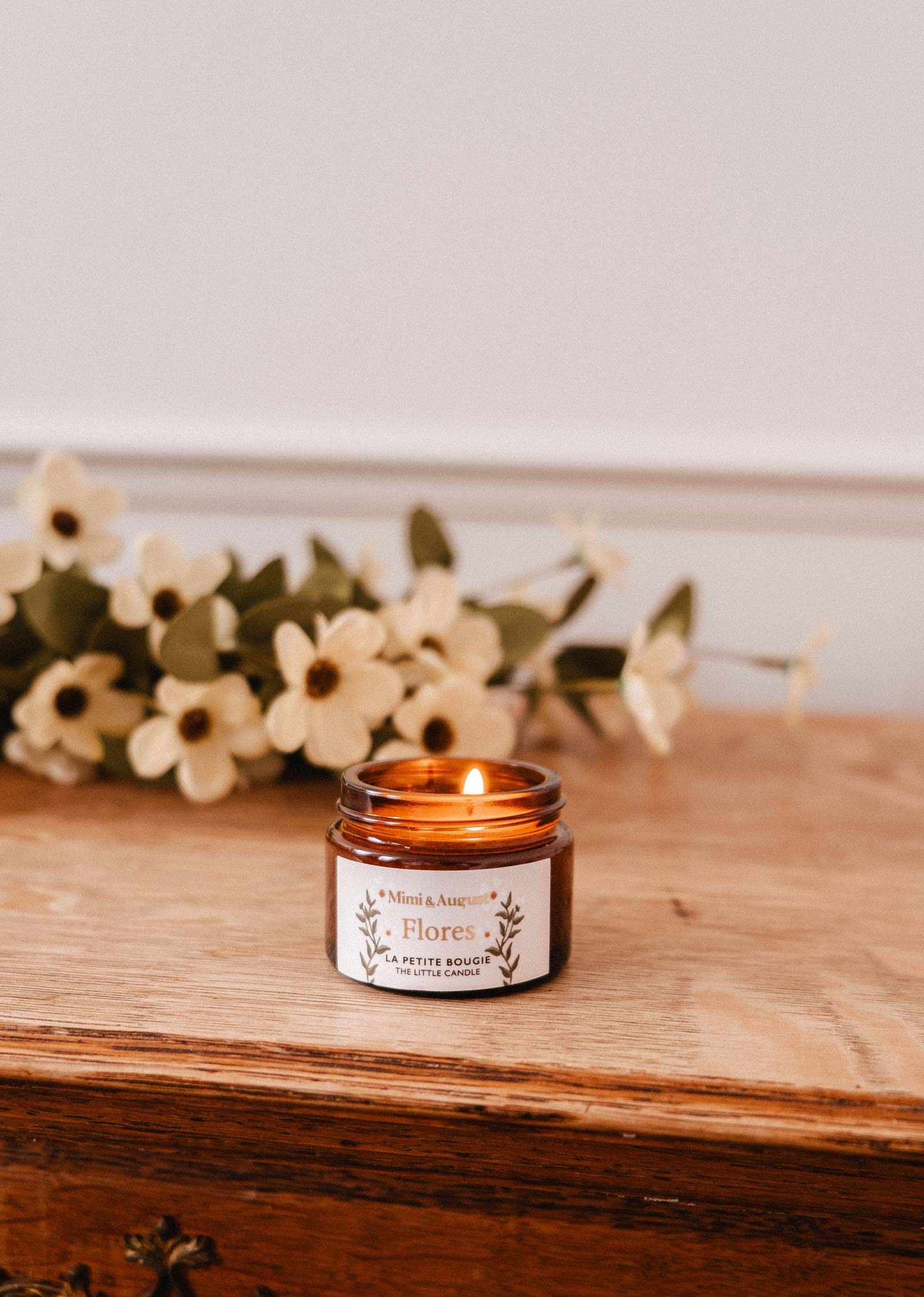 A 2 oz Flores soy candle by Mimi & August, in a small amber jar, is lit on a wooden surface with white flowers in the background. Hand-poured in Montreal.