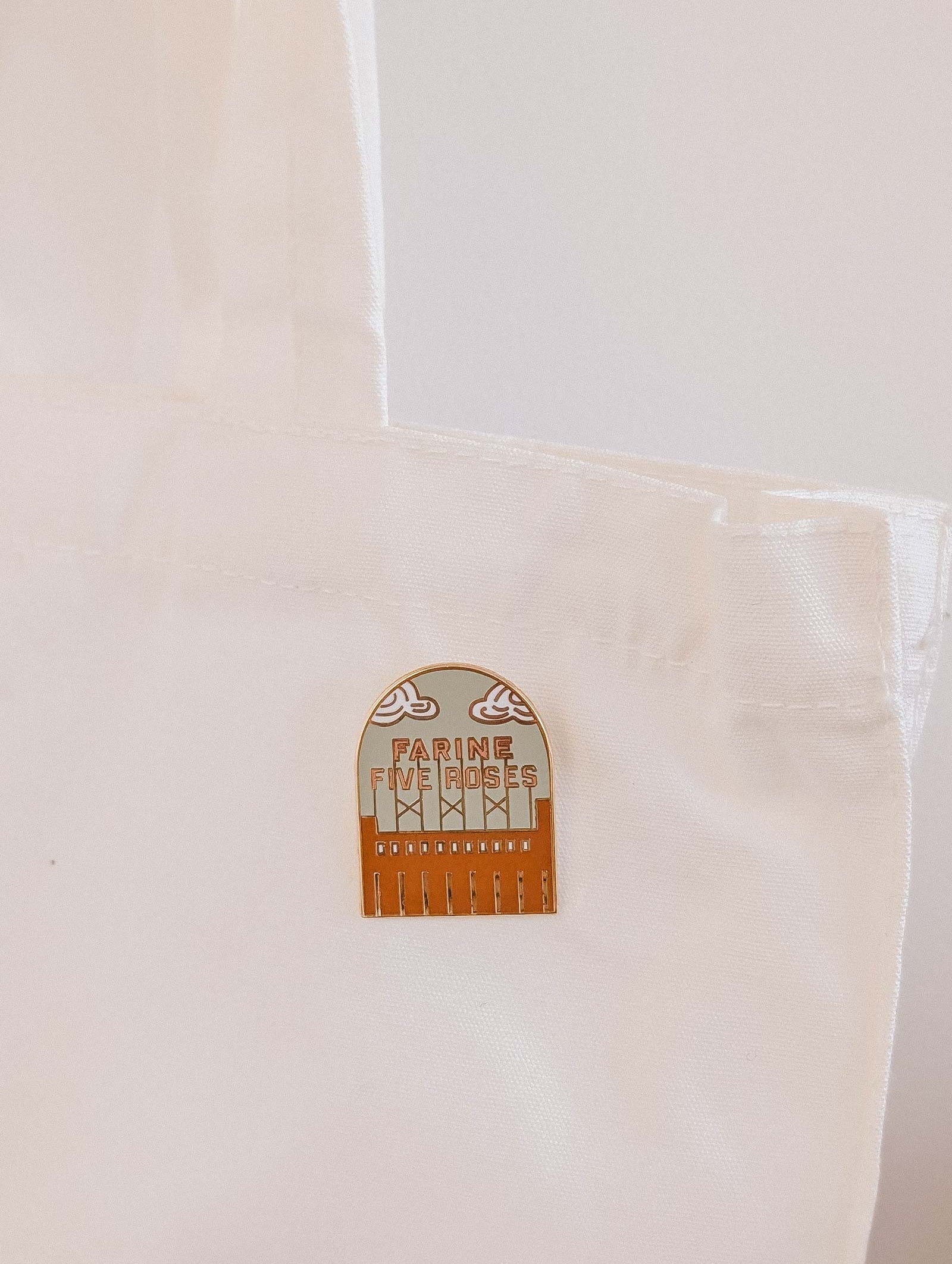 The Flour & Roses Factory Enamel Pin by Mimi & August is attached to the handle of a white fabric tote bag, adding a retro Montreal-inspired accent.