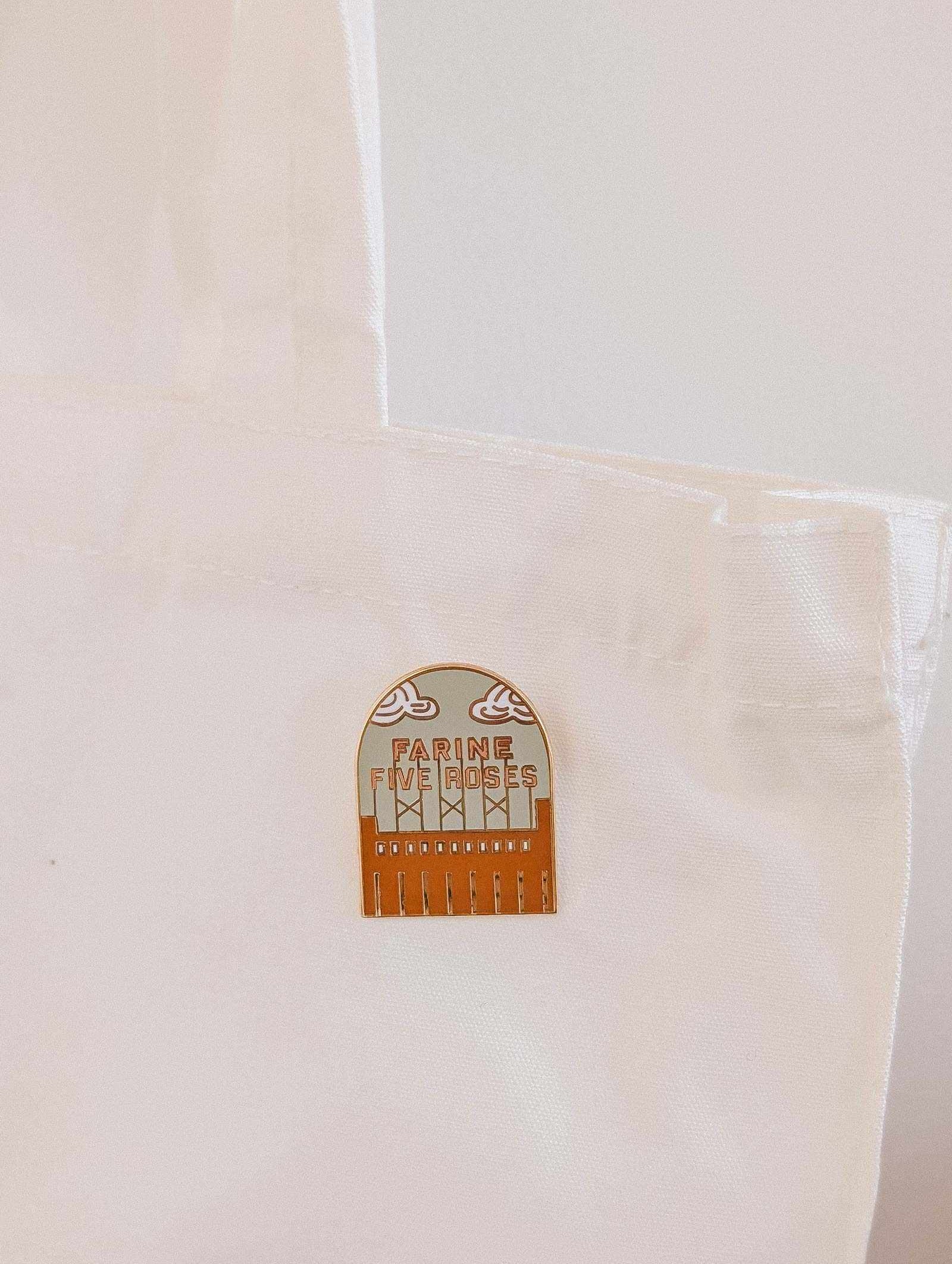 The Flour & Roses Factory Enamel Pin by Mimi & August is attached to the handle of a white fabric tote bag, adding a retro Montreal-inspired accent.