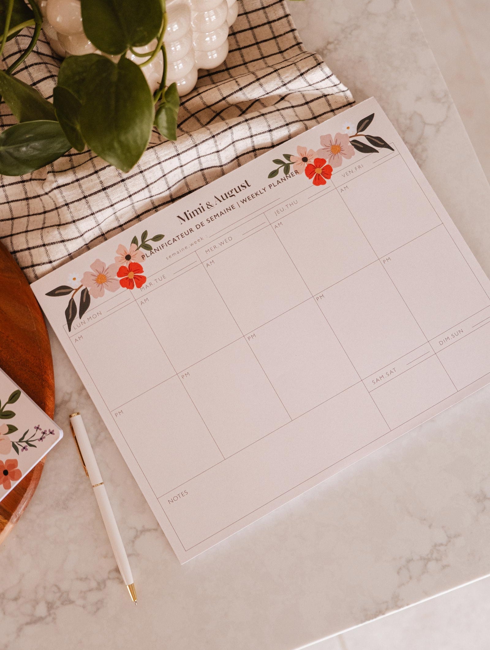 An eco-friendly Flower Bomb - Weekly Planner by Mimi & August on a table next to a plant and a pen.