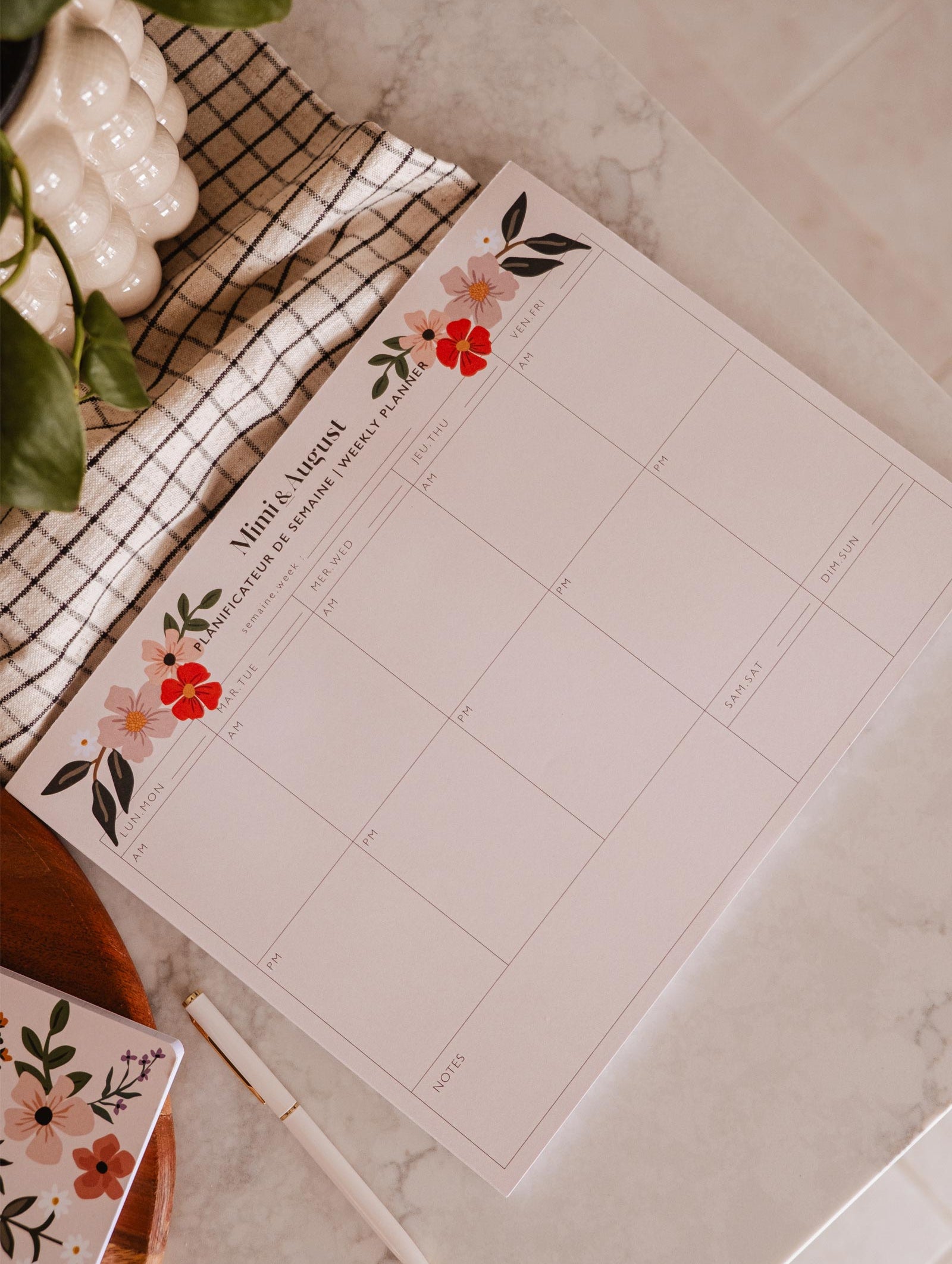 An eco-friendly Flower Bomb - Weekly Planner adorned with beautiful flowers, placed on a table alongside a potted plant. (Brand: Mimi & August)