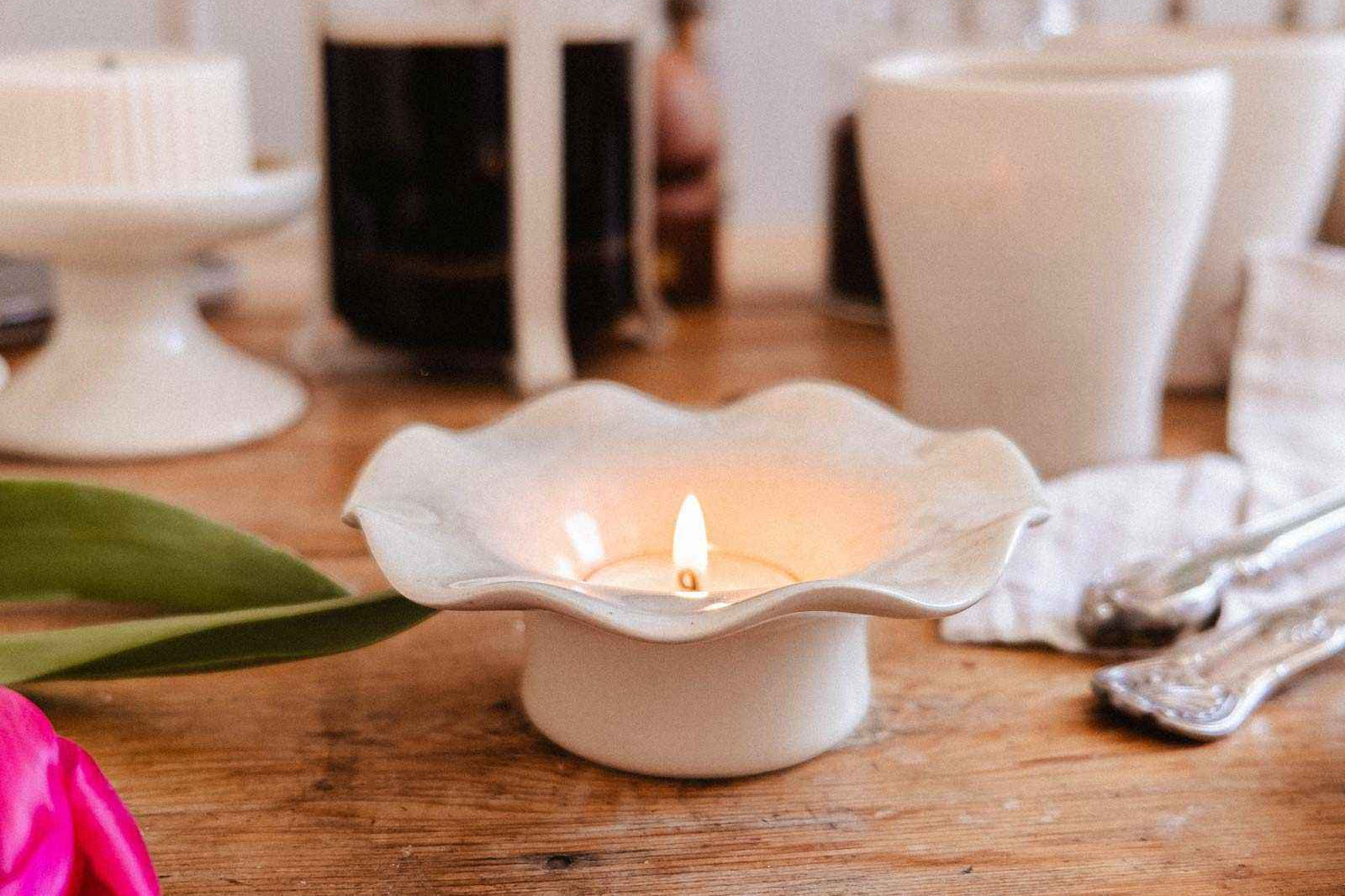 A lit candle in the Mimi & August Fluted Ceramic Tealight Holder sits on a wooden table with a French press, cups, cutlery, and pink tulips.