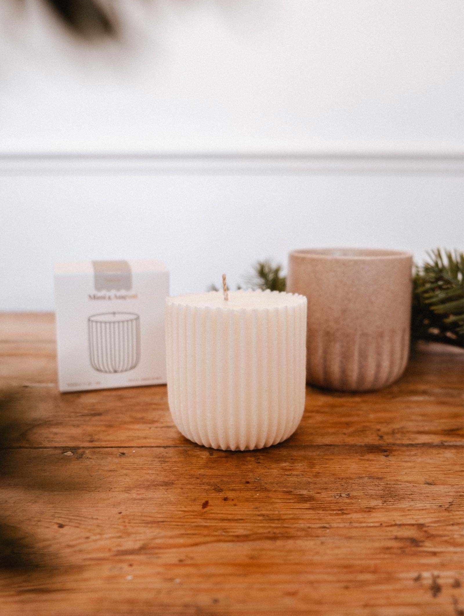 La Recharge de Bougie Forêt de Mimi & August, une bougie de soya blanche cannelée, repose sur une table en bois près d'une tasse brune, d'une petite boîte et de branches de conifère.