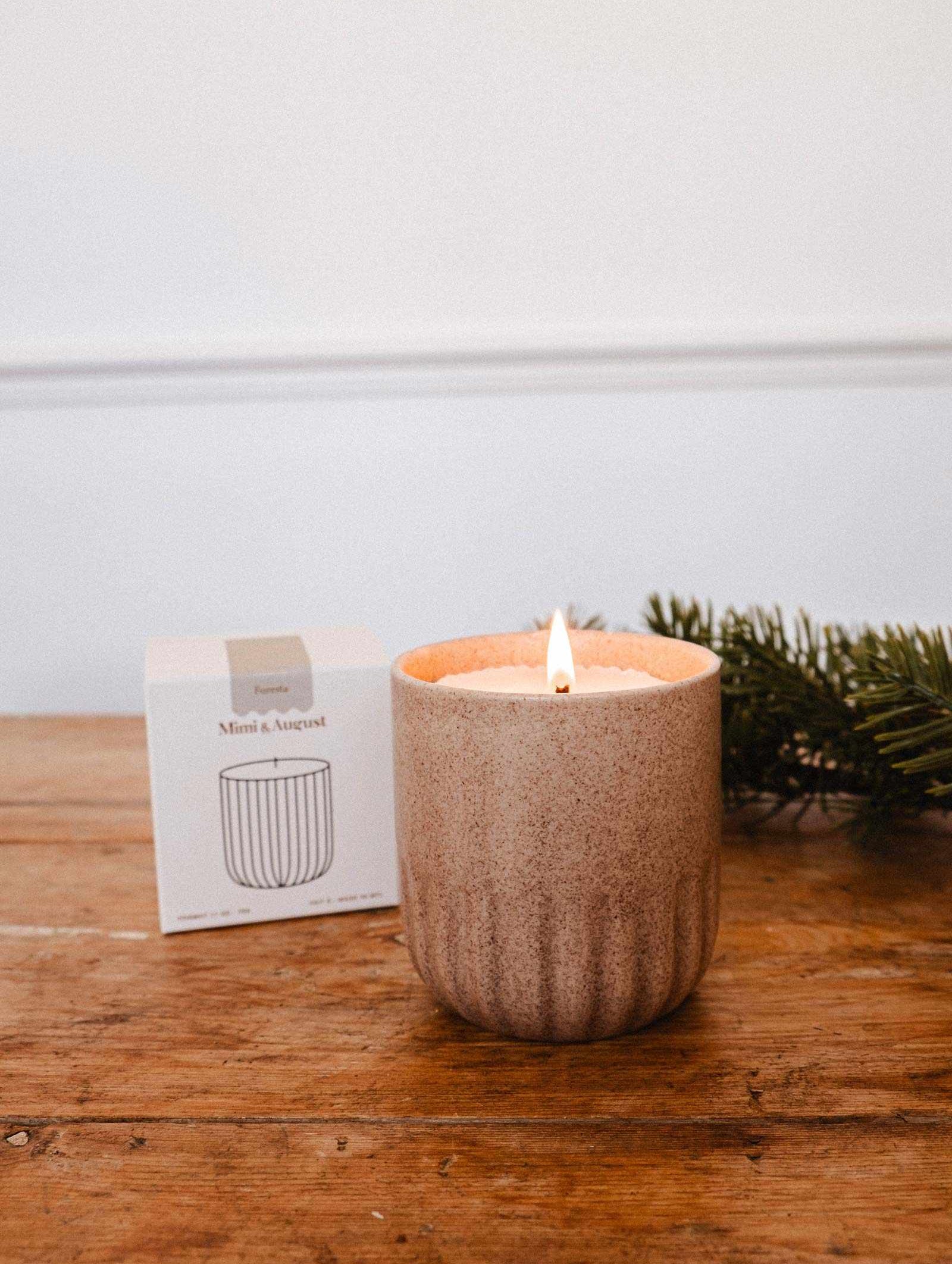 La Recharge de bougie Foresta de Mimi & August, fabriquée en cire de soya, repose dans un contenant texturé beige sur une surface en bois à côté de sa boîte et de verdure, libérant un subtil parfum de sapin dans l'air.