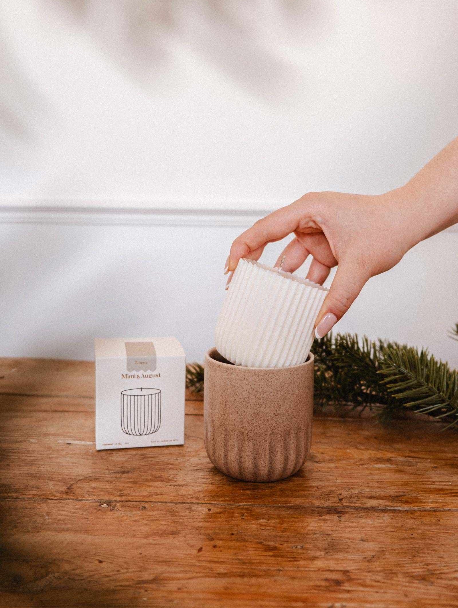 Une main dépose la Recharge de Bougie Forêt de Mimi & August, une bougie de soya blanche cannelée, dans un bougeoir beige en céramique sur une table en bois à côté d'une petite boîte et d'une branche de pin.