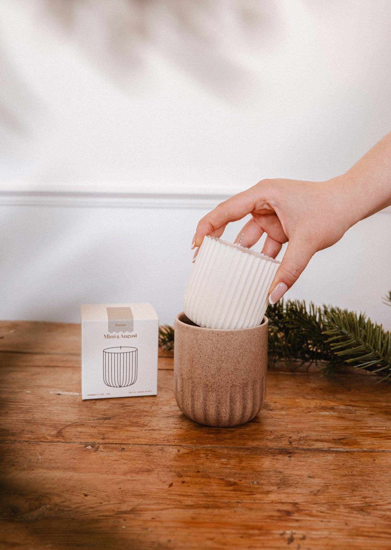 Une main dépose la Recharge de Bougie Forêt de Mimi & August, une bougie de soya blanche cannelée, dans un bougeoir beige en céramique sur une table en bois à côté d'une petite boîte et d'une branche de pin.