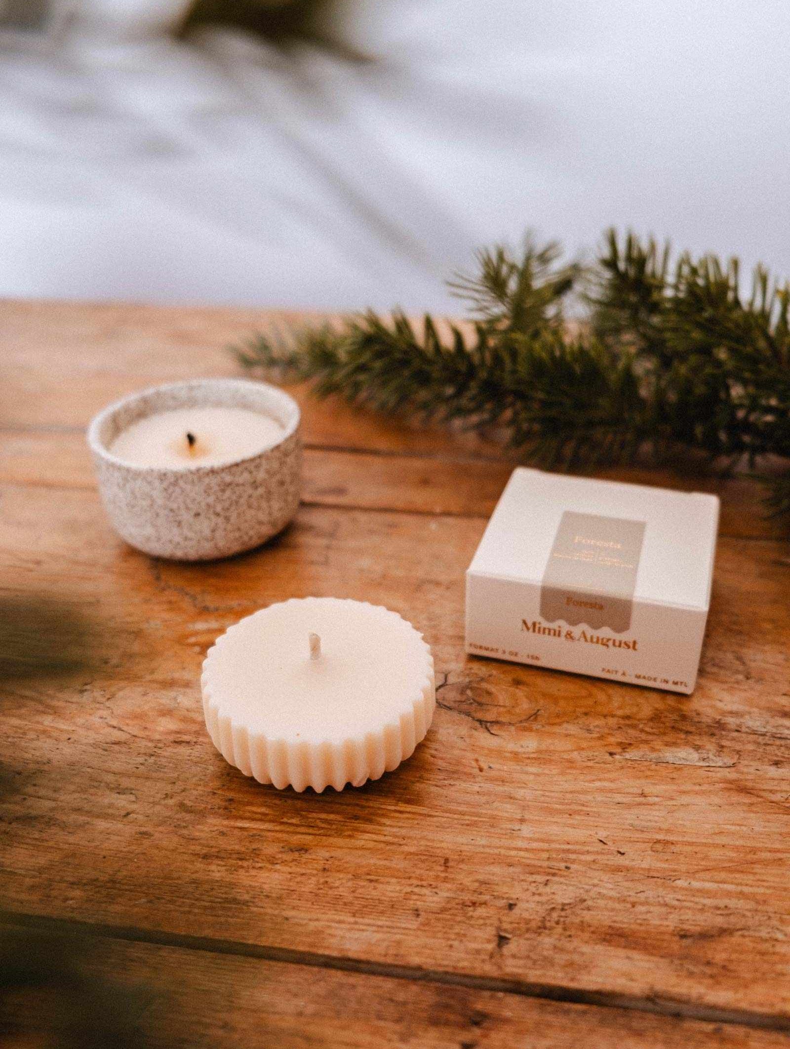 La Recharge de Bougie Forêt de Mimi & August, une bougie de soya blanche dans un plat texturé aux rebords cannelés avec une boîte étiquetée, est présentée sur du bois avec des branches de pin, capturant l'arôme frais de sapin.