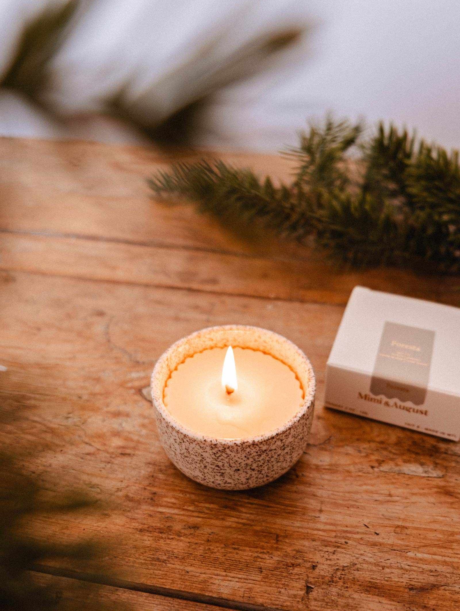 Recharge de Bougie Forêt de Mimi & August, avec son subtil parfum de sapin, repose dans un bougeoir en céramique moucheté sur du bois à côté d'une boîte fermée et de branches de pin, emplissant l'air de fragrance.