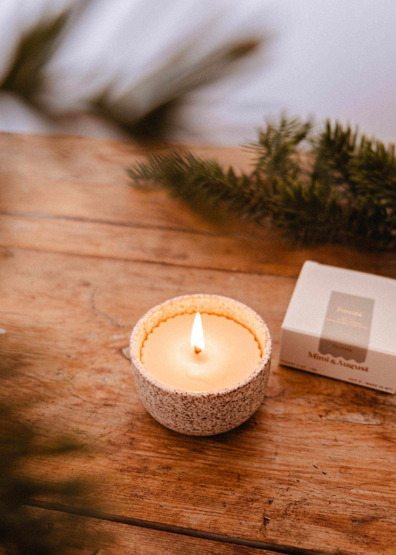 La Recharge de bougie Foresta de Mimi & August, avec son subtil parfum de sapin, repose dans un bougeoir en céramique moucheté sur du bois à côté d'une boîte fermée et de branches de pin, emplissant l'air de fragrance.