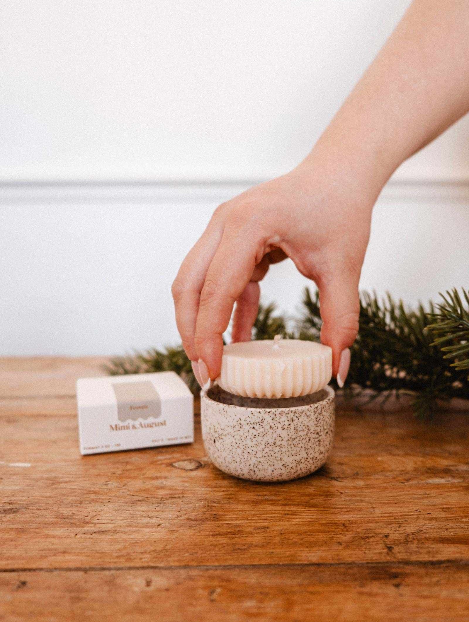 Une main dépose un rond de savon sur un contenant en céramique moucheté, avec la Recharge de bougie Foresta de Mimi & August et une branche de pin sur une surface en bois en arrière-plan.