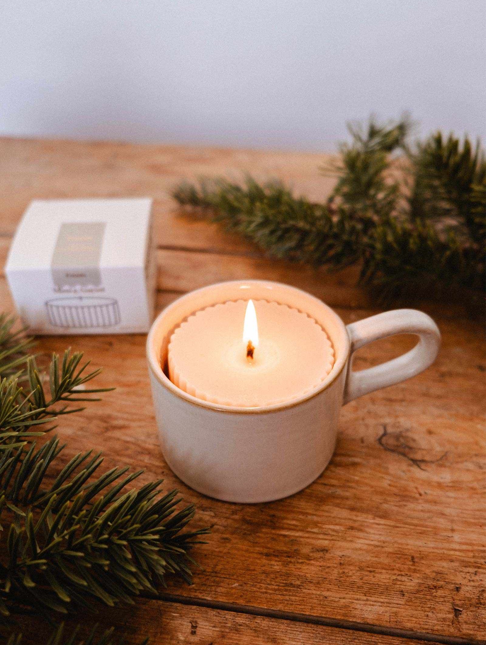 La Recharge de bougie Foresta de Mimi & August, une bougie de soya au doux arôme de sapin, repose dans une tasse en céramique sur du bois parmi des branches de pin et une boîte blanche.