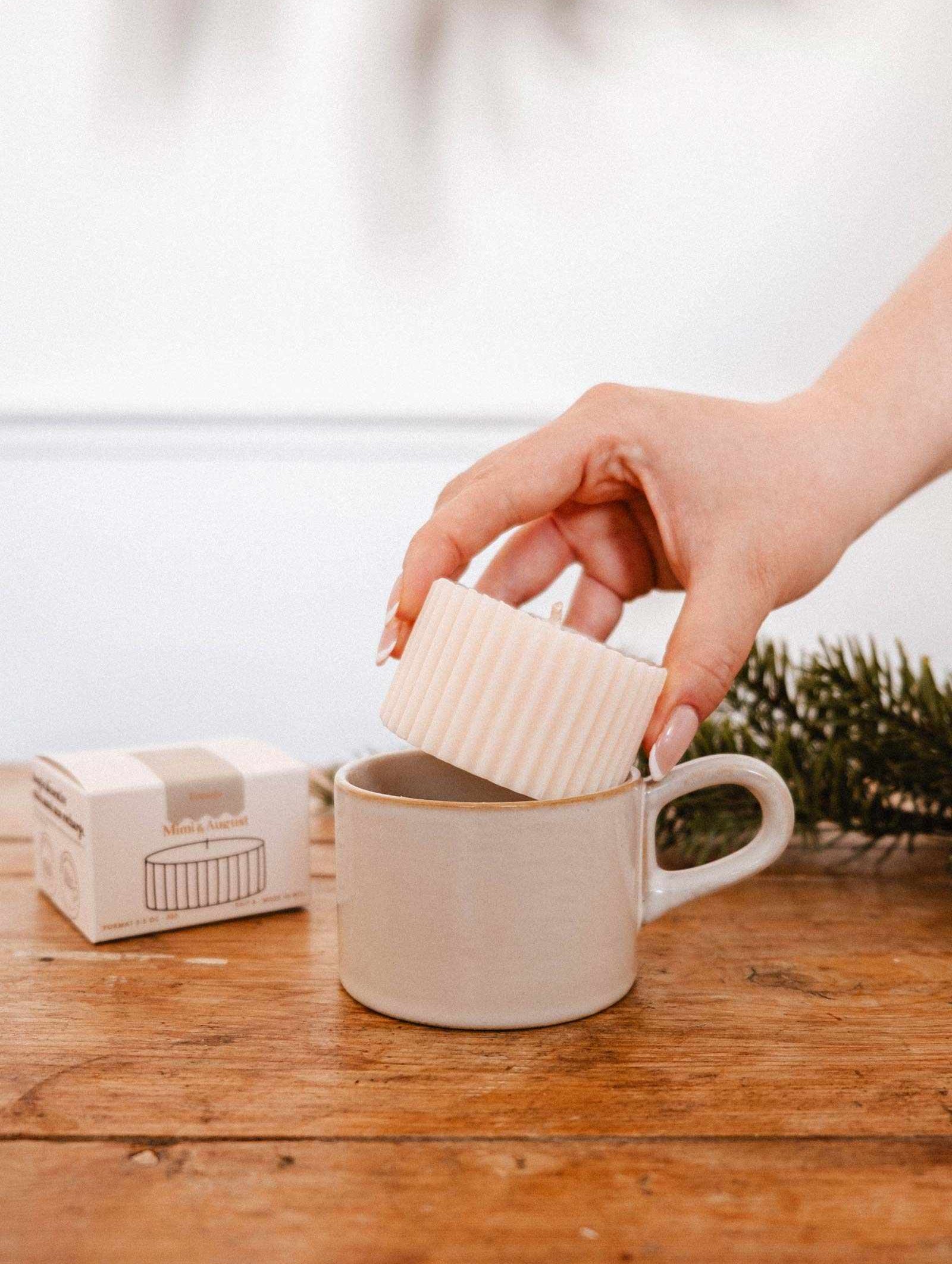 Une main dépose la Recharge de Bougie Forêt de Mimi & August dans une tasse beige en céramique sur une table en bois, avec sa boîte et une branche de pin en arrière-plan.