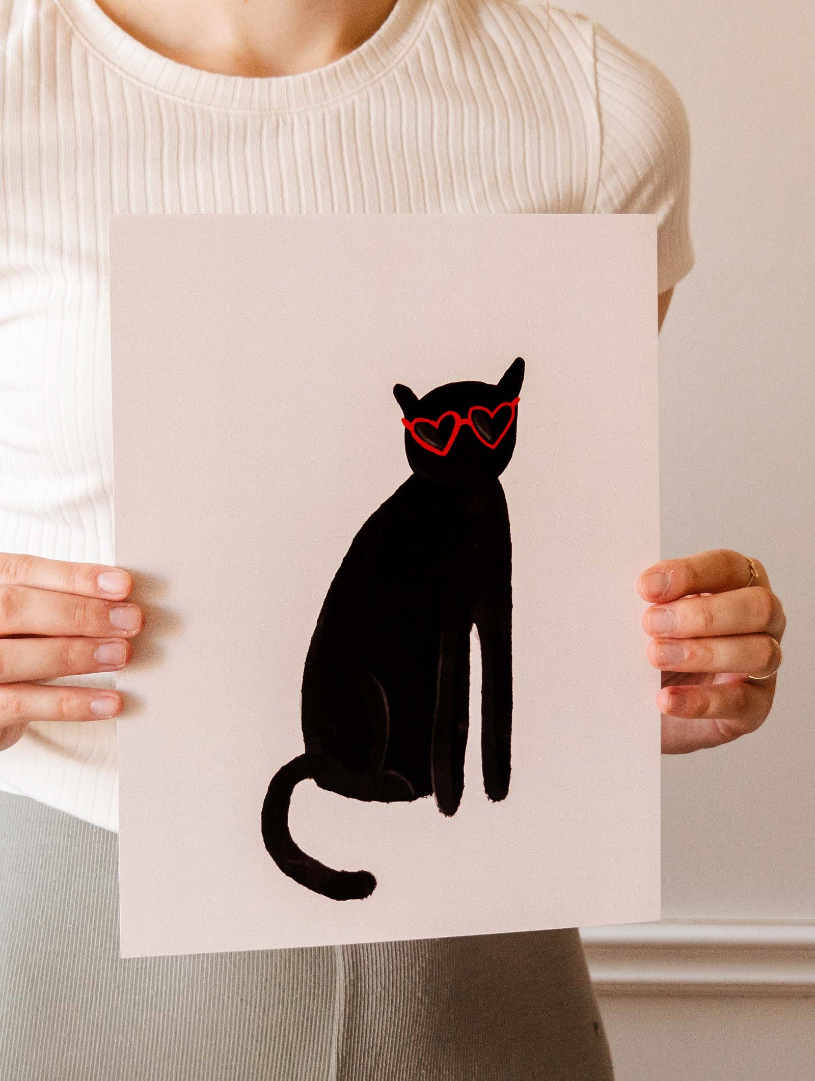 A woman holding up a Mimi & August Heart Black Cat Art Print wearing heart-shaped sunglasses.