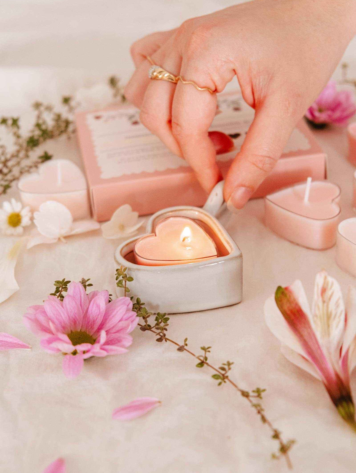 A hand lights a Little Hearts – Tealight Candle from Mimi & August in a ceramic holder, surrounded by pink flowers and romantic decor, with more heart-shaped tealights arranged on a white surface.