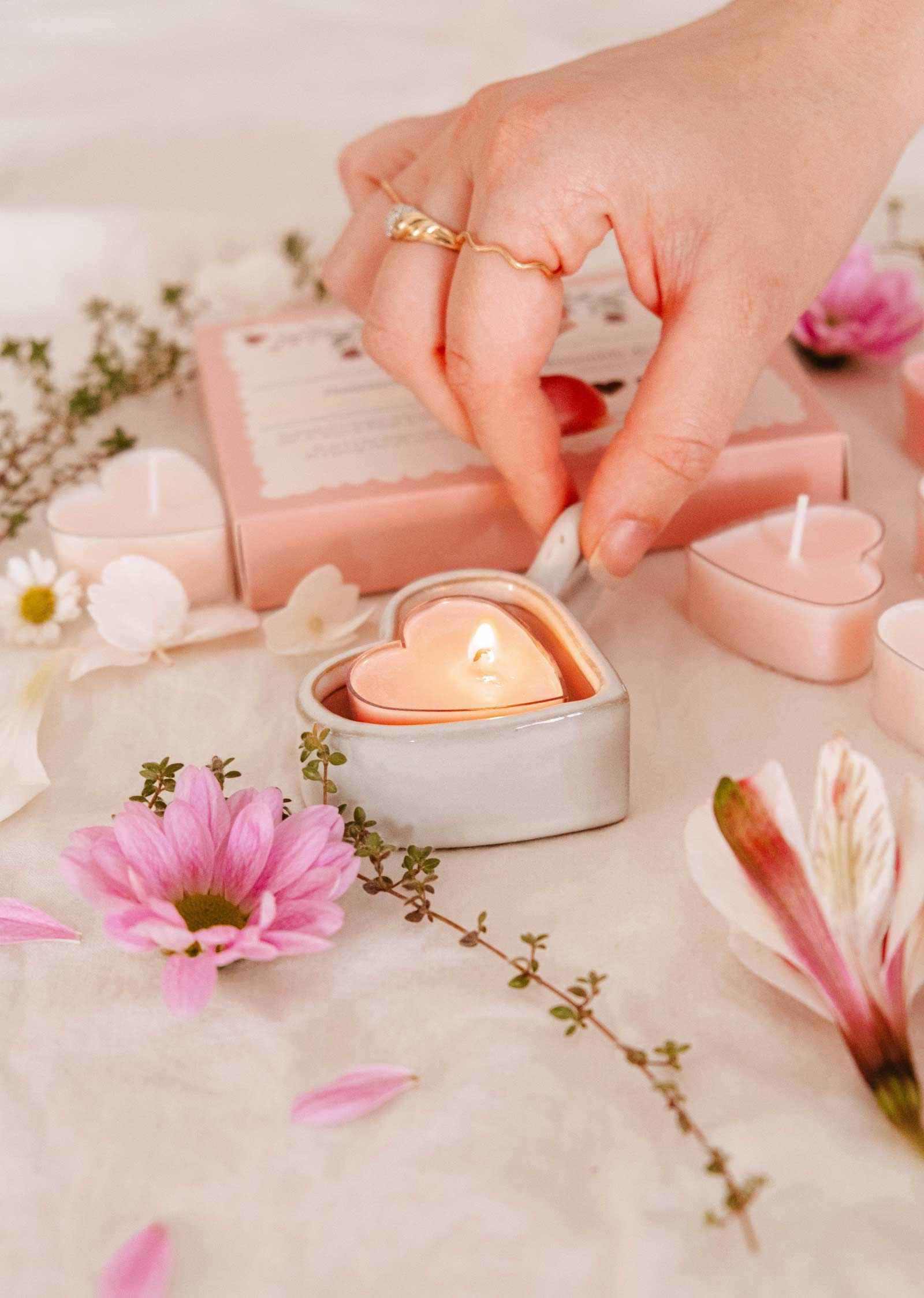 A hand lights a Little Hearts – Tealight Candle from Mimi & August in a ceramic holder, surrounded by pink flowers and romantic decor, with more heart-shaped tealights arranged on a white surface.