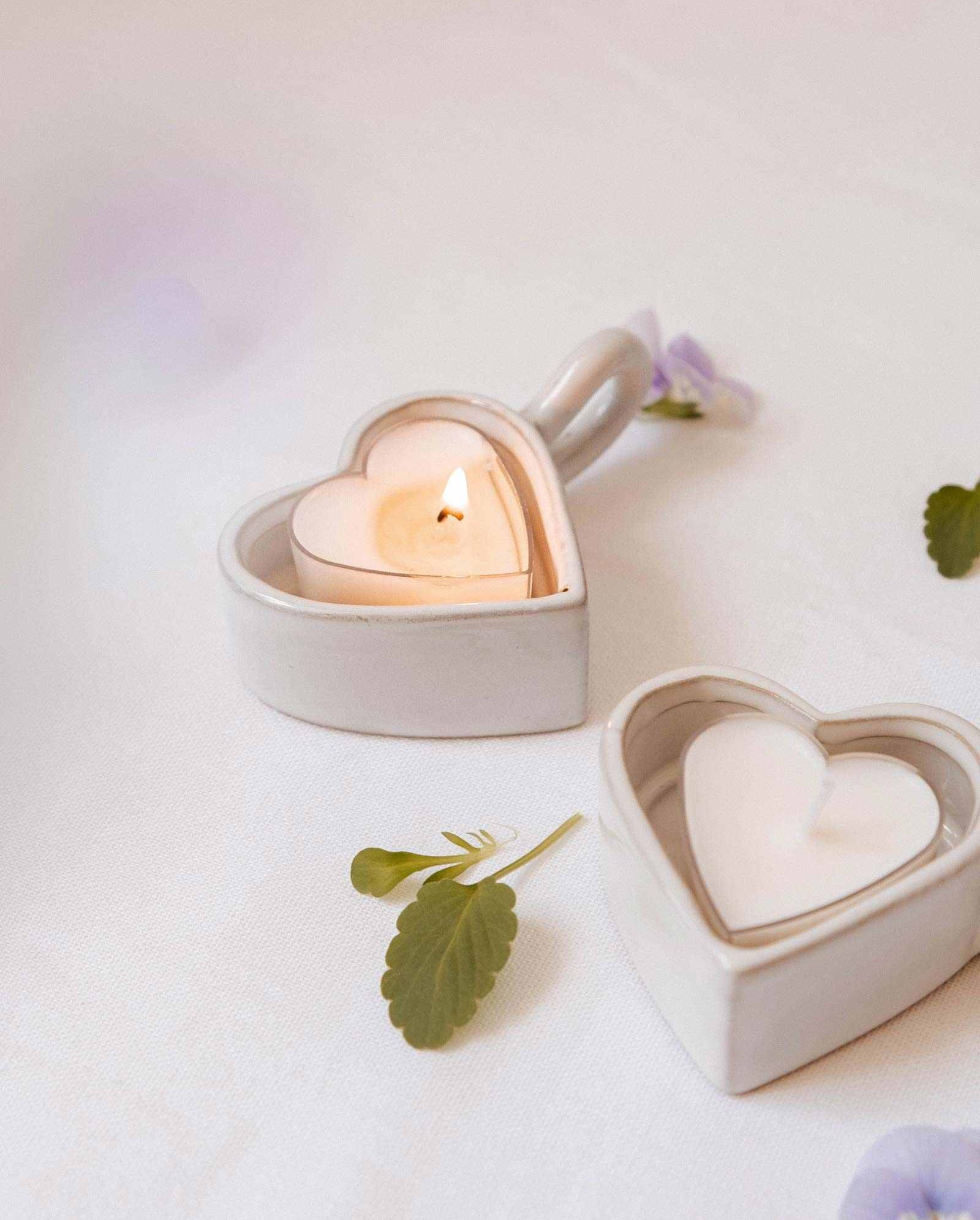 Two Mimi & August Heart & Glow Tealight Holders, one lit and one unlit, sit on a white surface with delicate flowers and leaves scattered nearby, highlighting their heart-shaped ceramic design.
