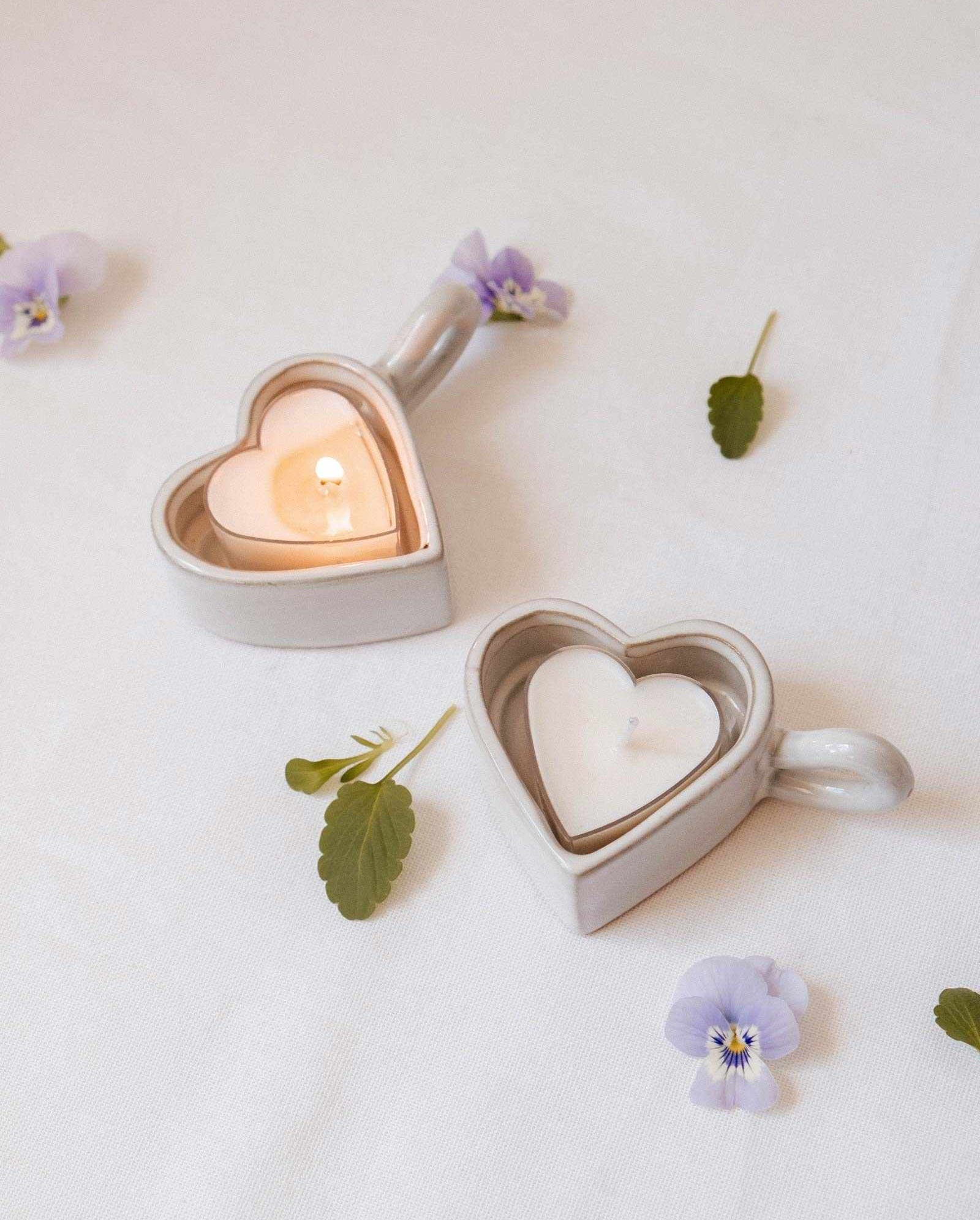 Two Heart & Glow Tealight Holders by Mimi & August, one with a lit and one with an unlit tealight, sit on a white cloth decorated with scattered purple flowers and green leaves.