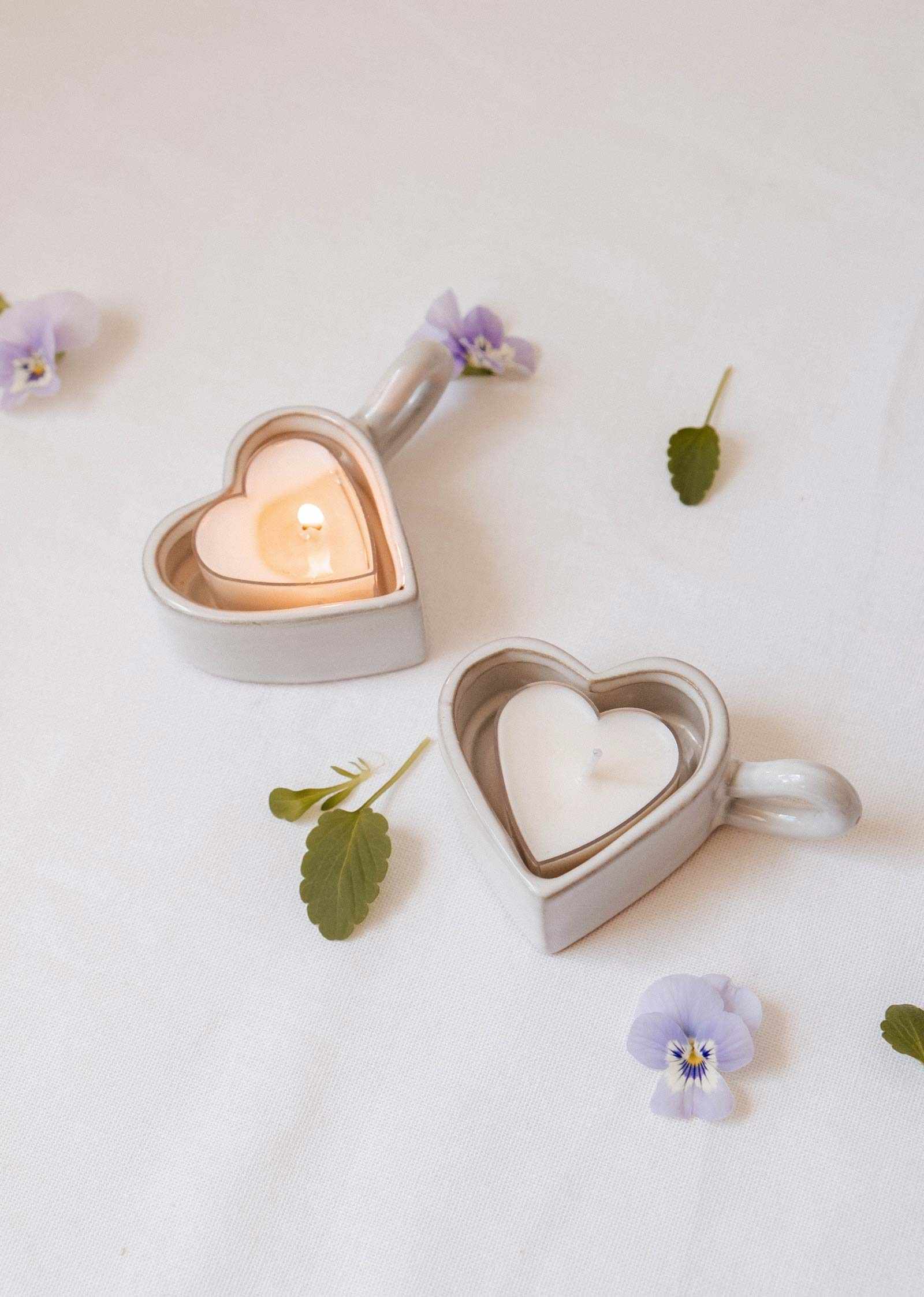 Two Heart & Glow Tealight Holders by Mimi & August, one with a lit and one with an unlit tealight, sit on a white cloth decorated with scattered purple flowers and green leaves.