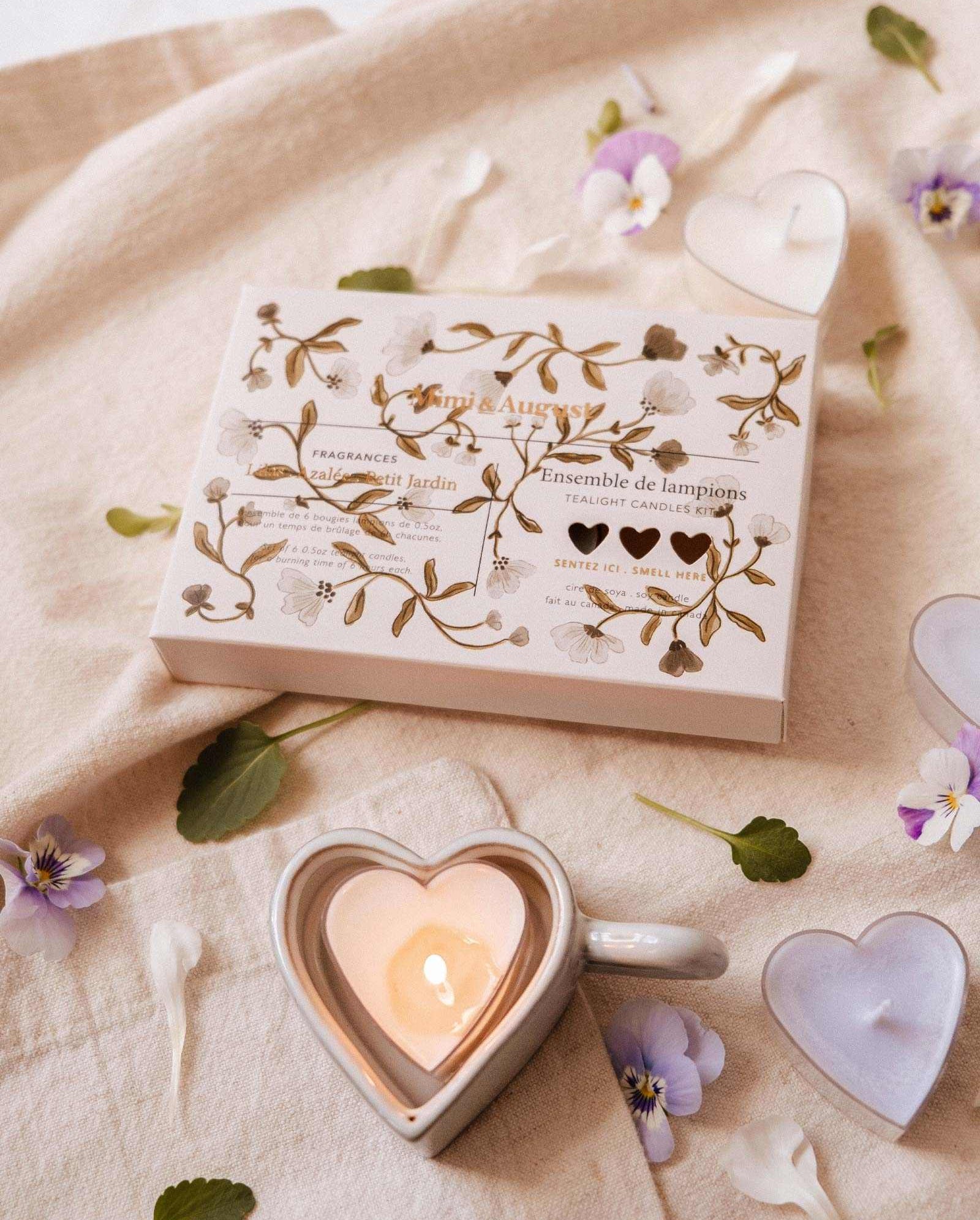 A Heart & Glow Tealight Holder by Mimi & August rests on a beige cloth, surrounded by Blush Hearts Candle Tealights, heart-shaped candles, flower petals, and greenery.