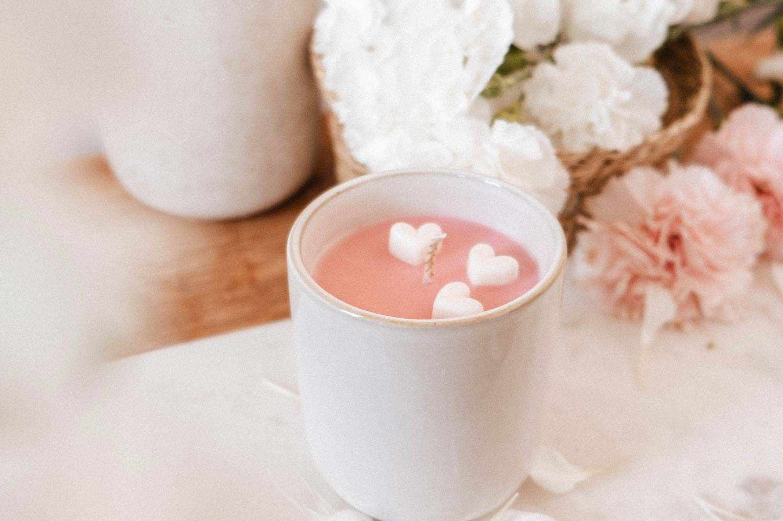 The Special Edition Candle by Mimi & August, a pink limited edition candle in a white cup with heart-shaped wax pieces, sits on marble among white and pink flowers.