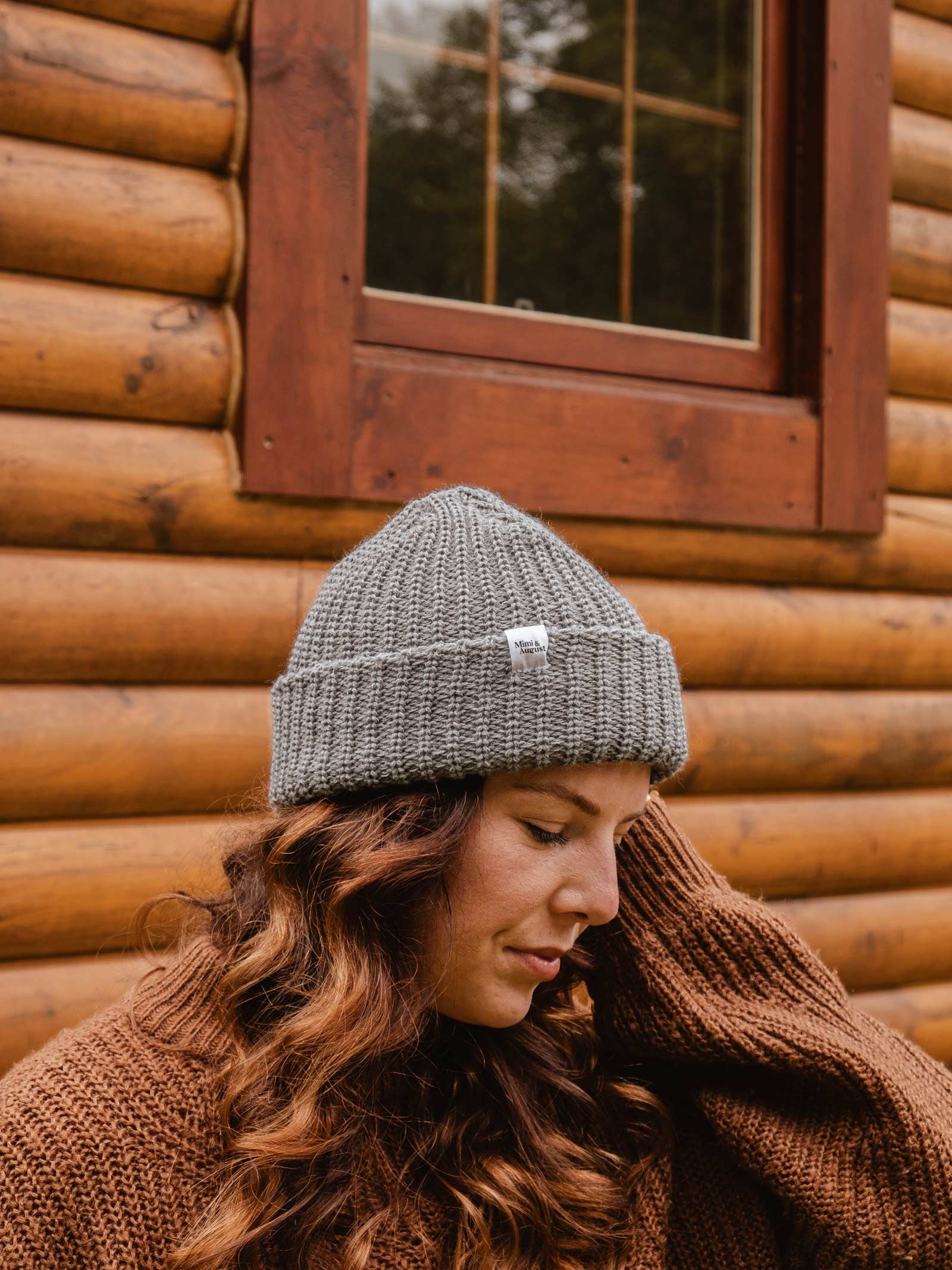 A person with long, wavy hair, wearing a Heather Grey Chunky Beanie from Mimi & August and a brown sweater, stands in front of a wooden cabin wall and window.