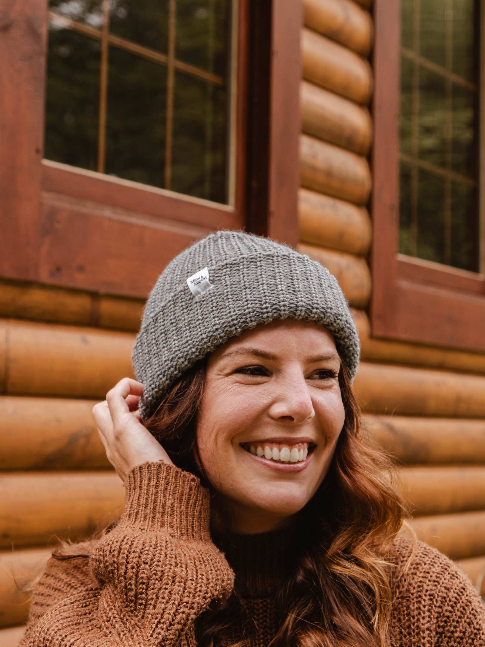 A woman wearing a Mimi & August Heather Grey Chunky Beanie and a brown sweater smiles in front of a wooden cabin.