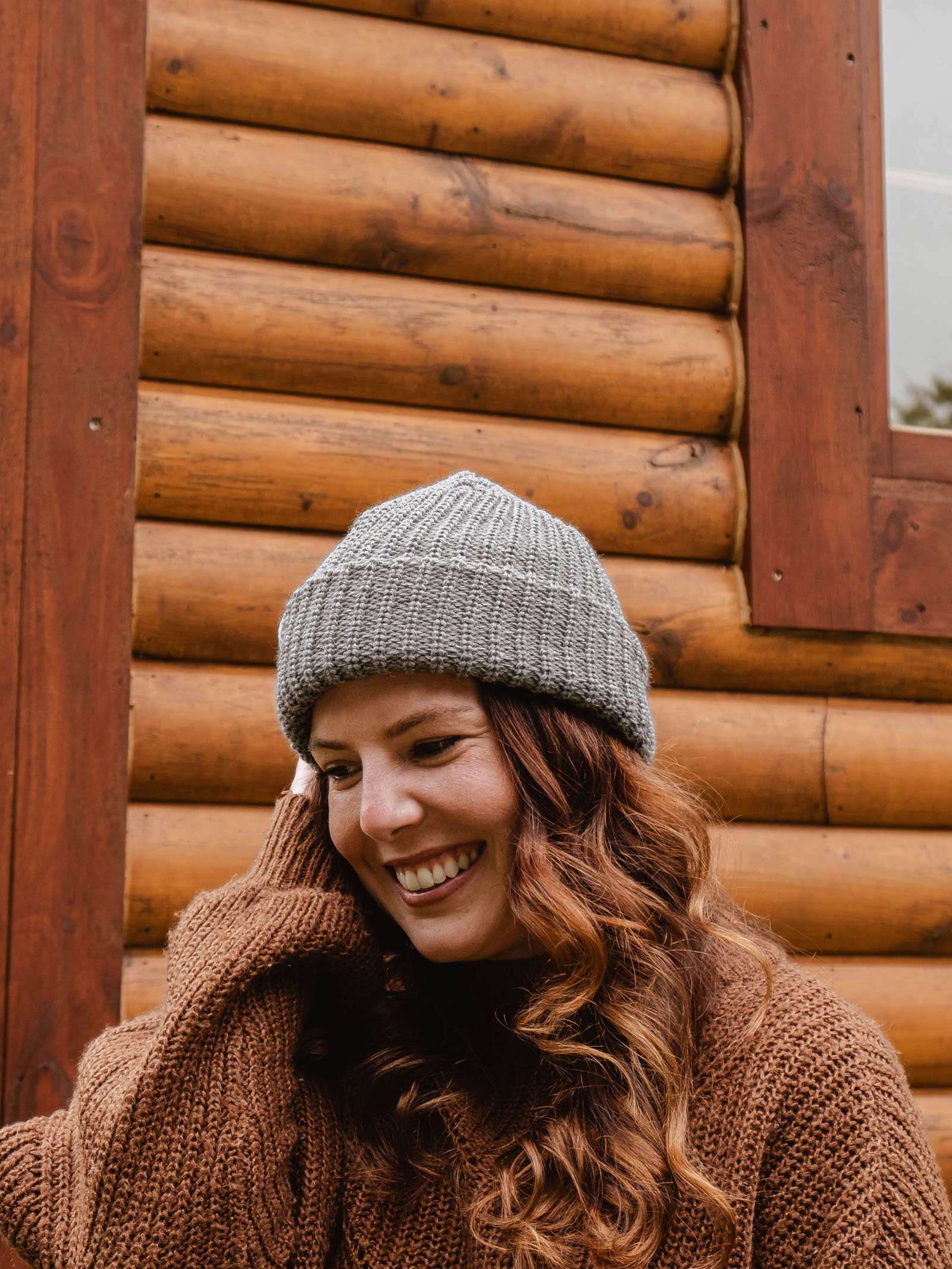 A person wearing a Heather Grey Chunky Beanie by Mimi & August and a sweater stands in front of a log cabin with a smile.