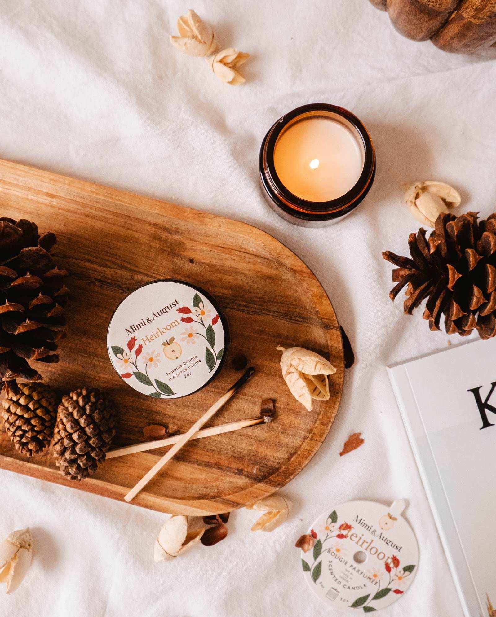 A wooden tray adorned with pinecones, the Heirloom reusable candle from Mimi & August emitting a rich cedar fragrance, and small decorative items on a white surface, beside a partially visible magazine or book.