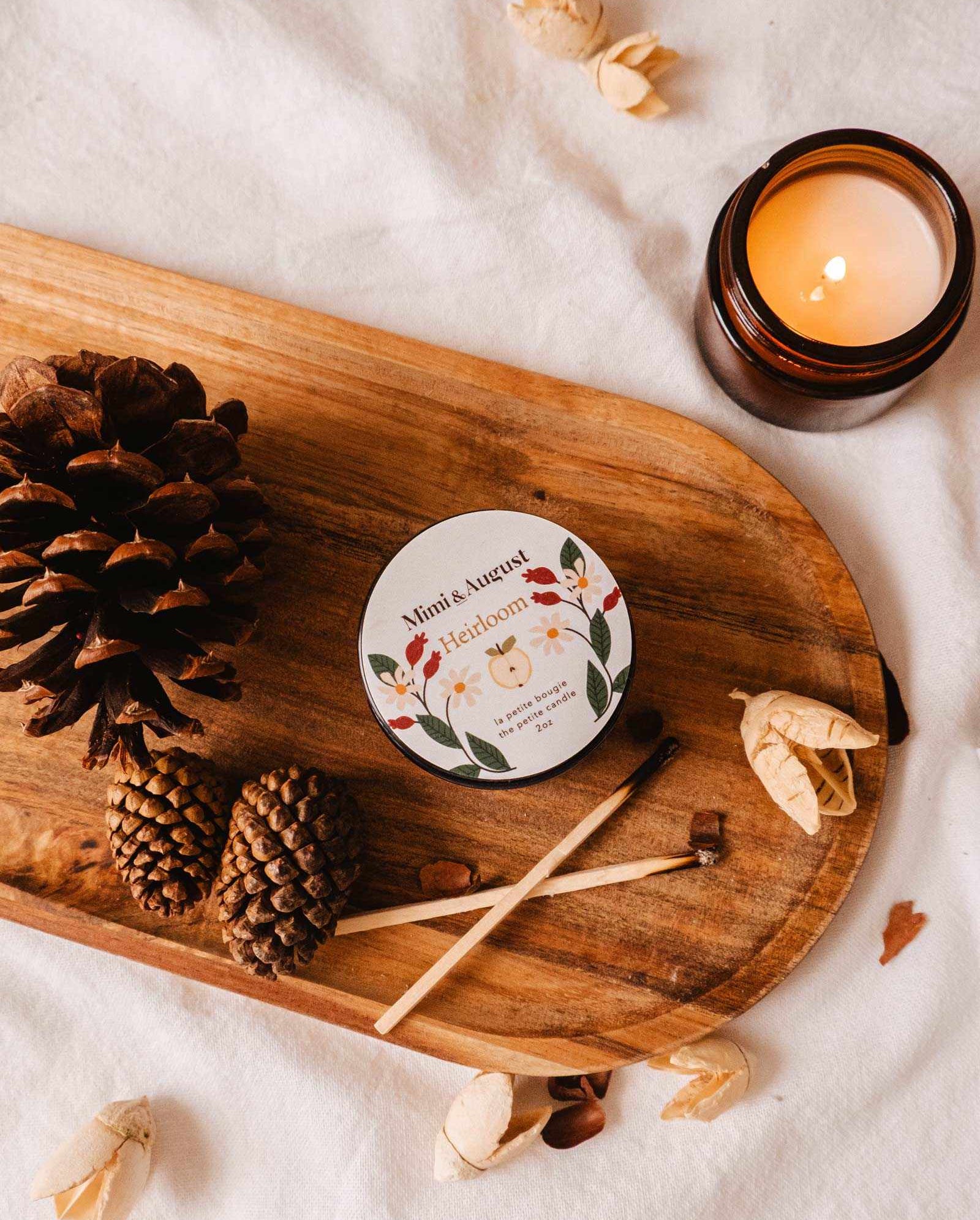A lit Heirloom - Reusable Candle by Mimi & August, a container labeled "Heirloom," pinecones, and matches are arranged on a wooden tray placed on a white fabric background, with the subtle scents of cedar and clove enhancing the cozy ambiance.