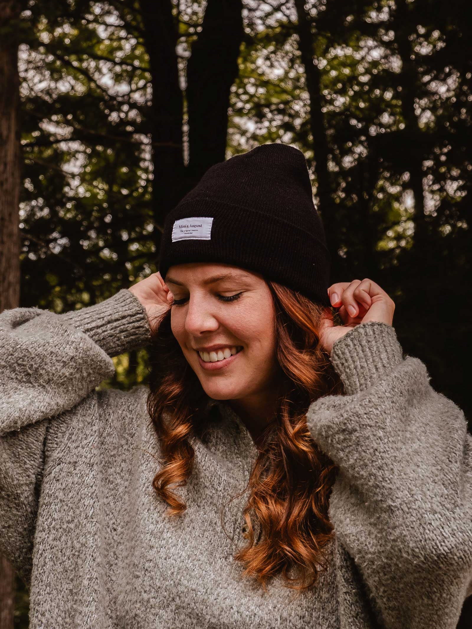 A person wearing a gray sweater and an Intemporel Black Beanie from Mimi & August smiles while standing in a forested area, showcasing their chic winter accessory.