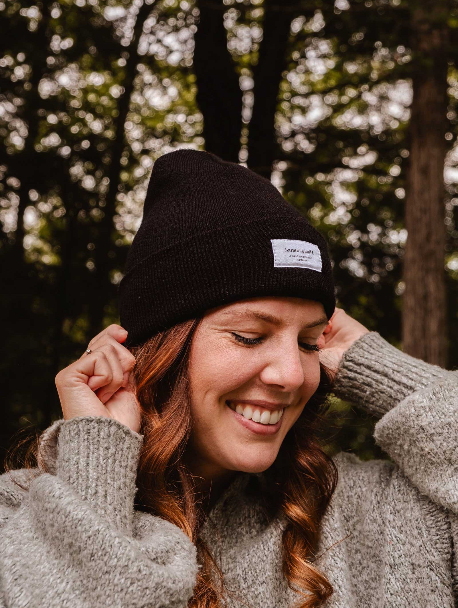 A person with long curly hair, wearing a Mimi & August Intemporel Black Beanie and a knitted sweater, smiles while standing outdoors with trees in the background.