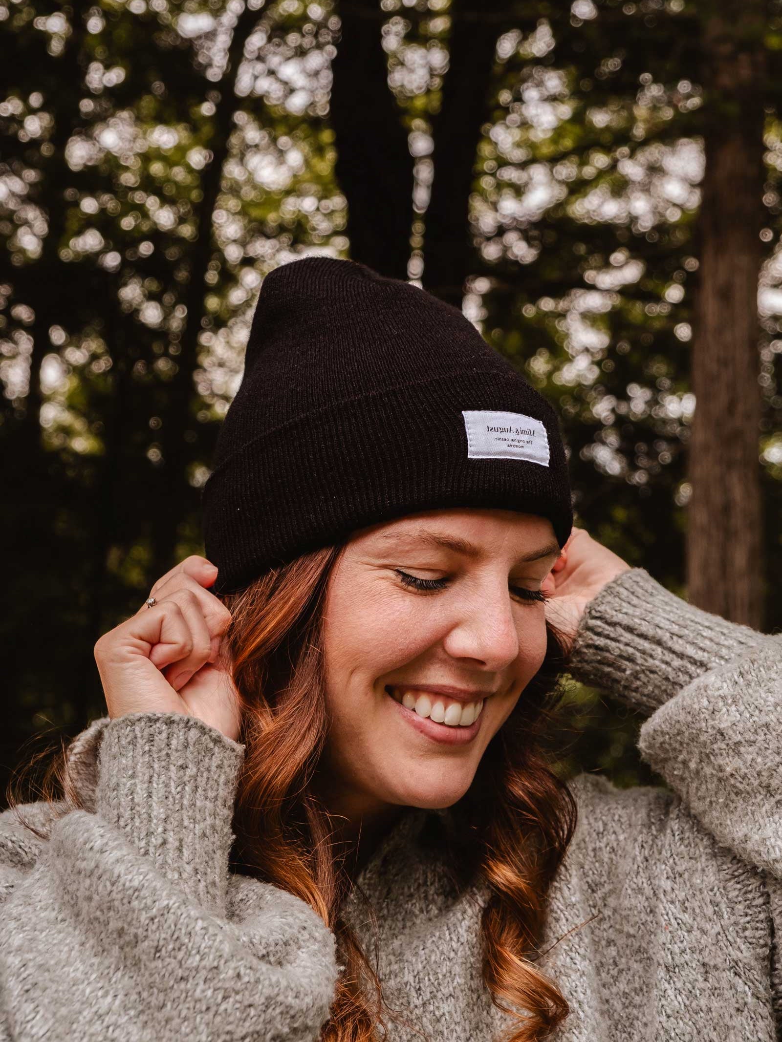 A person with long curly hair, wearing a Mimi & August Intemporel Black Beanie and a knitted sweater, smiles while standing outdoors with trees in the background.