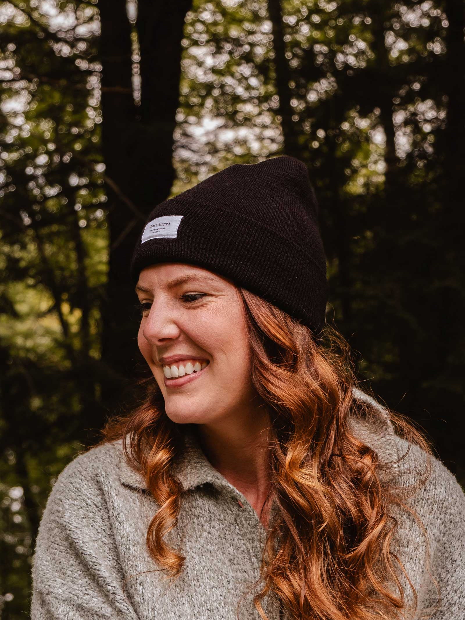 A woman with long, wavy hair wearing a Mimi & August Intemporel Black Beanie and a grey sweater smiles while outdoors. Trees are visible in the background, highlighting her stylish winter accessory.