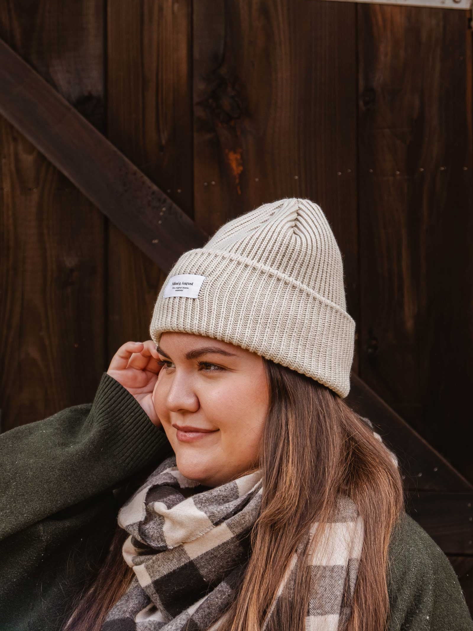 A person wearing a light-colored knit beanie, a green sweater, and a plaid scarf stands in front of a wooden background, showcasing their winter wardrobe featuring the unisex Ivory Snuggle Beanie by Mimi & August.
