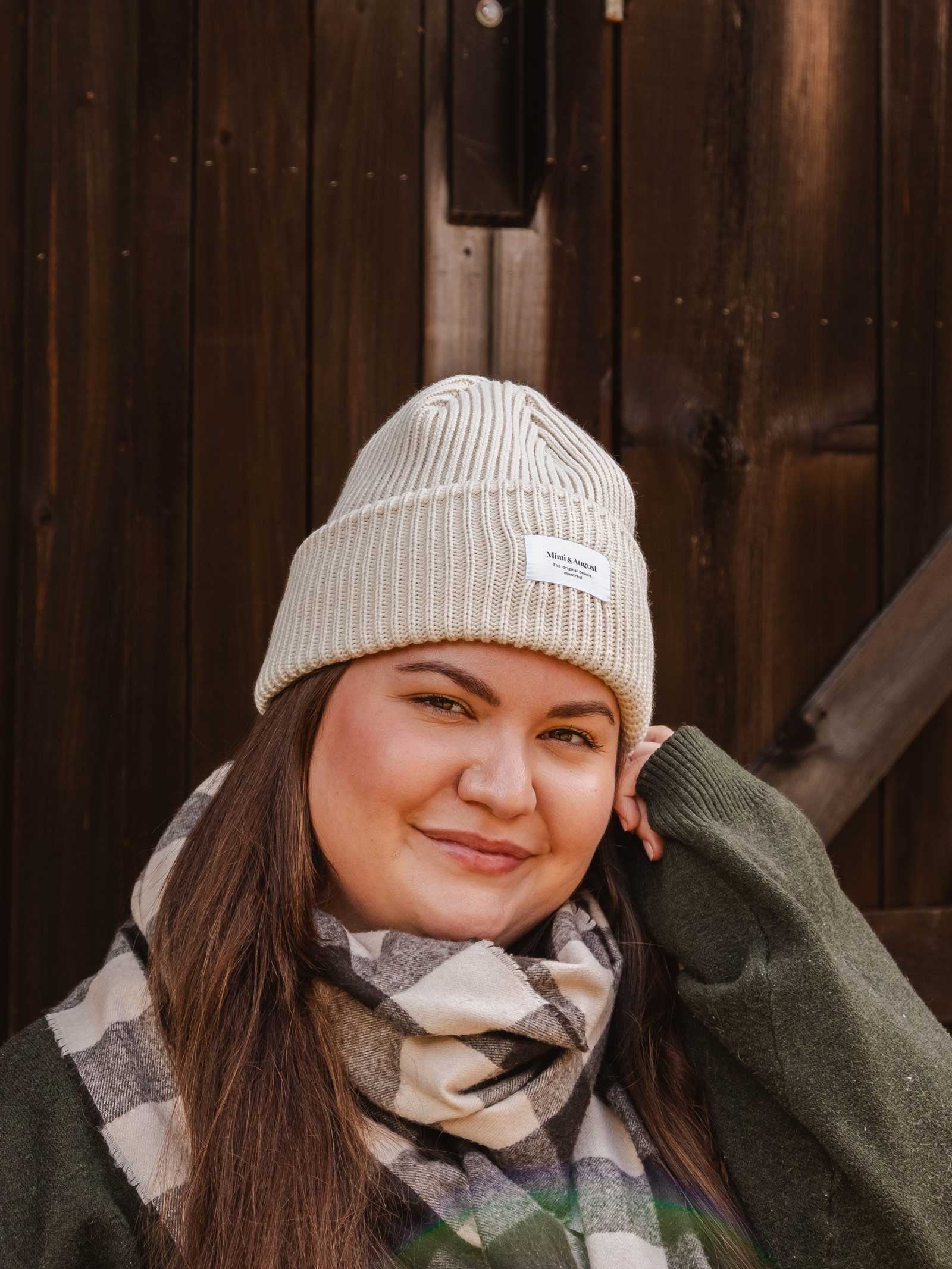 A person wearing a Mimi & August Ivory Snuggle Beanie, a green sweater, and a checkered scarf poses in front of a wooden background, showcasing their unisex acrylic knit to perfect their winter wardrobe.