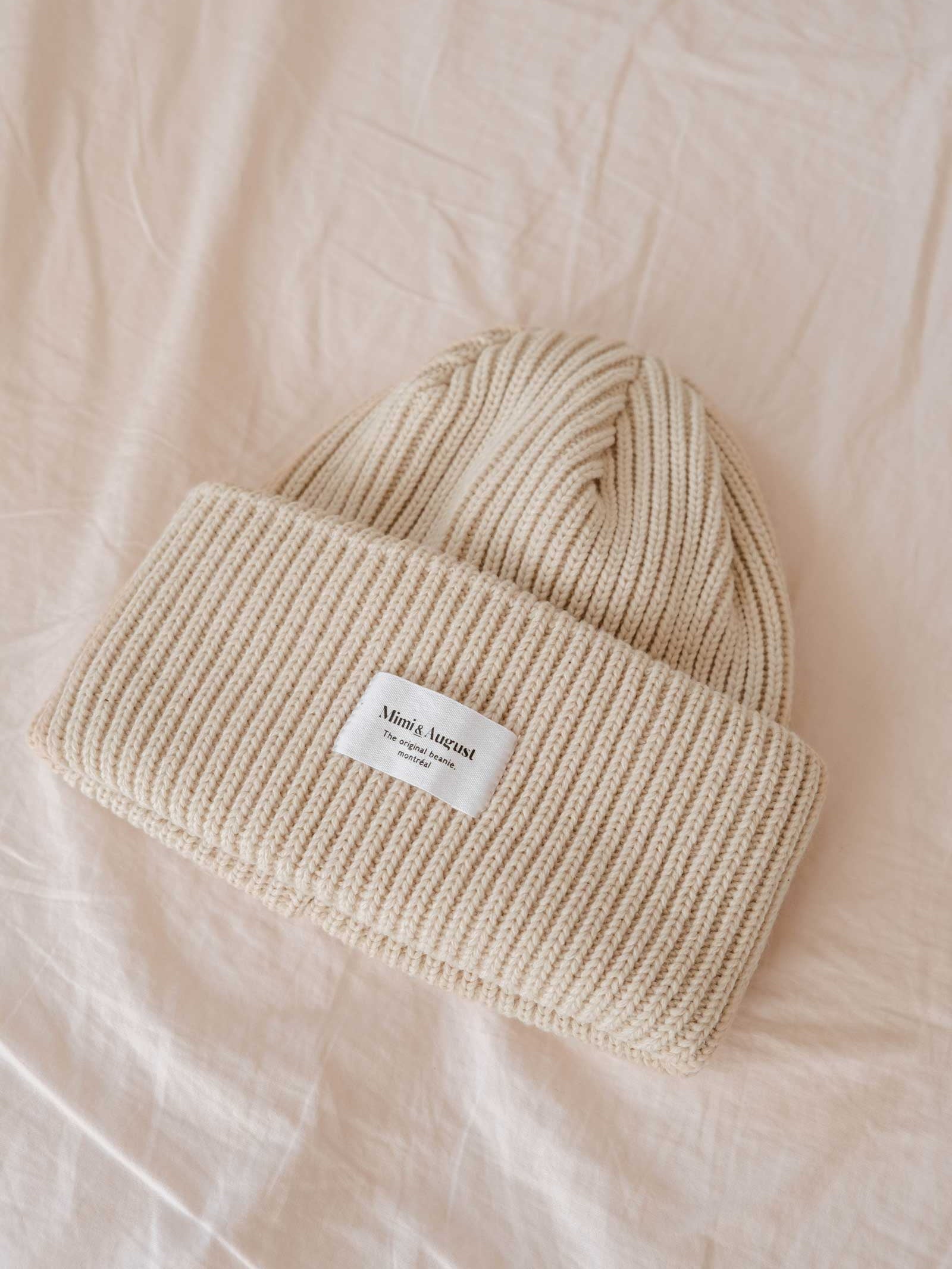 The Ivory Snuggle Beanie by Mimi & August, a beige ribbed accessory featuring a white logo patch, rests on a cream-colored fabric surface, making it an ideal and versatile addition to any winter wardrobe. This unisex acrylic knit ensures both style and warmth.