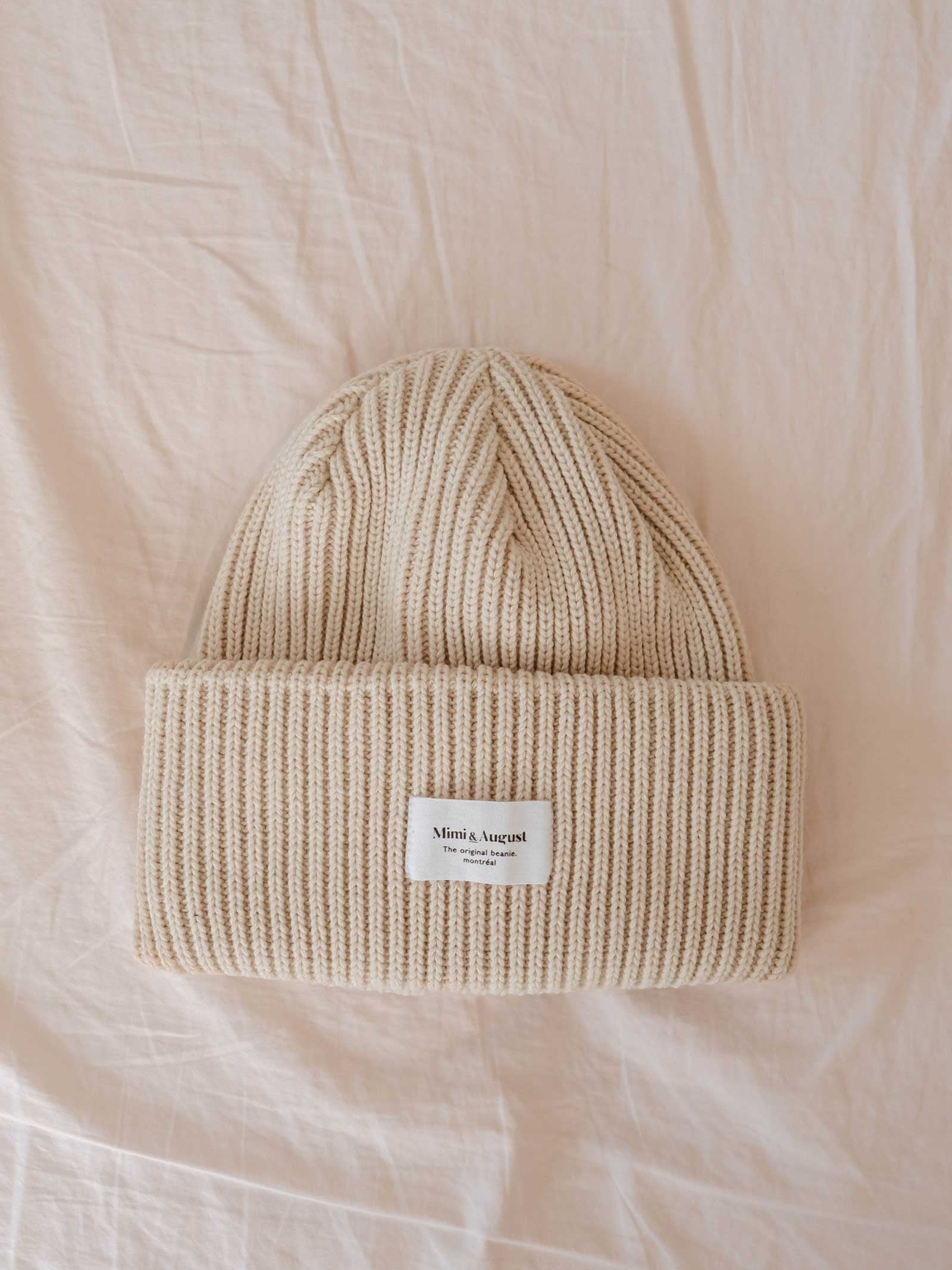 The Ivory Snuggle Beanie, a beige ribbed knit beanie featuring a white label with "Mimi & August," sits on a cream-colored fabric background. This unisex acrylic knit is a must-have for your winter wardrobe.