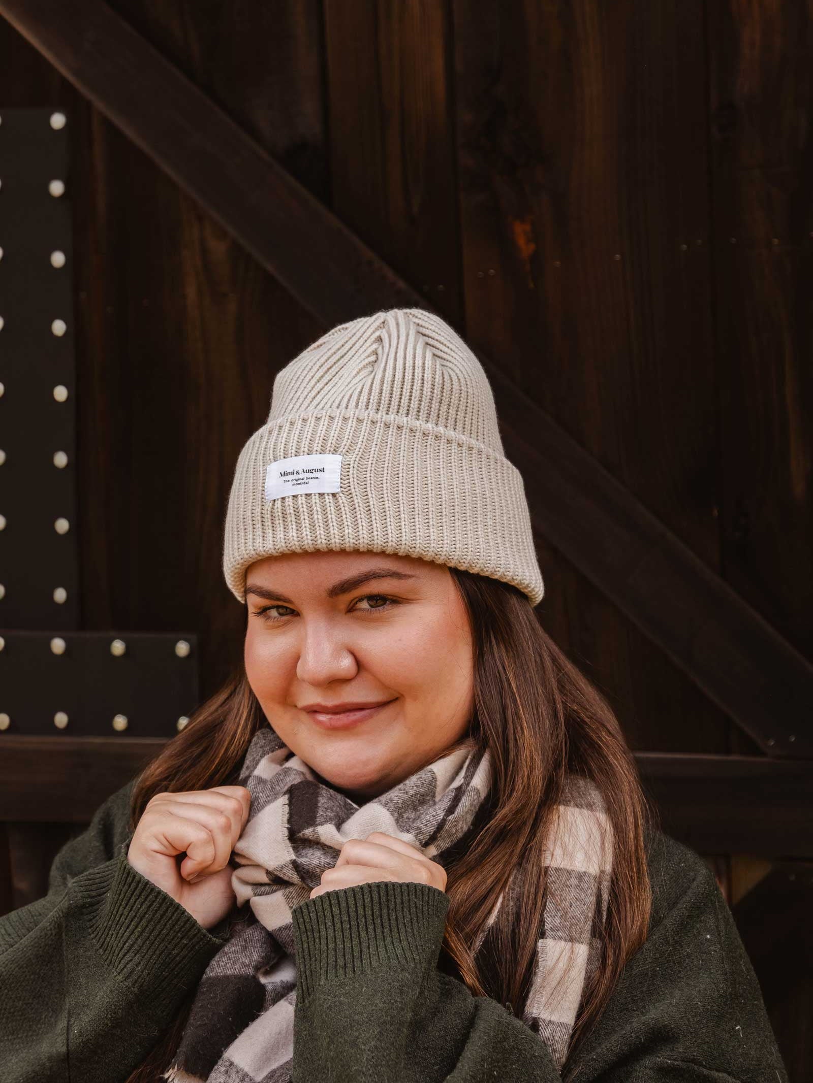 A person wearing a Mimi & August Ivory Snuggle Beanie and a checkered scarf stands in front of a dark wooden background, looking at the camera, showcasing their winter wardrobe.