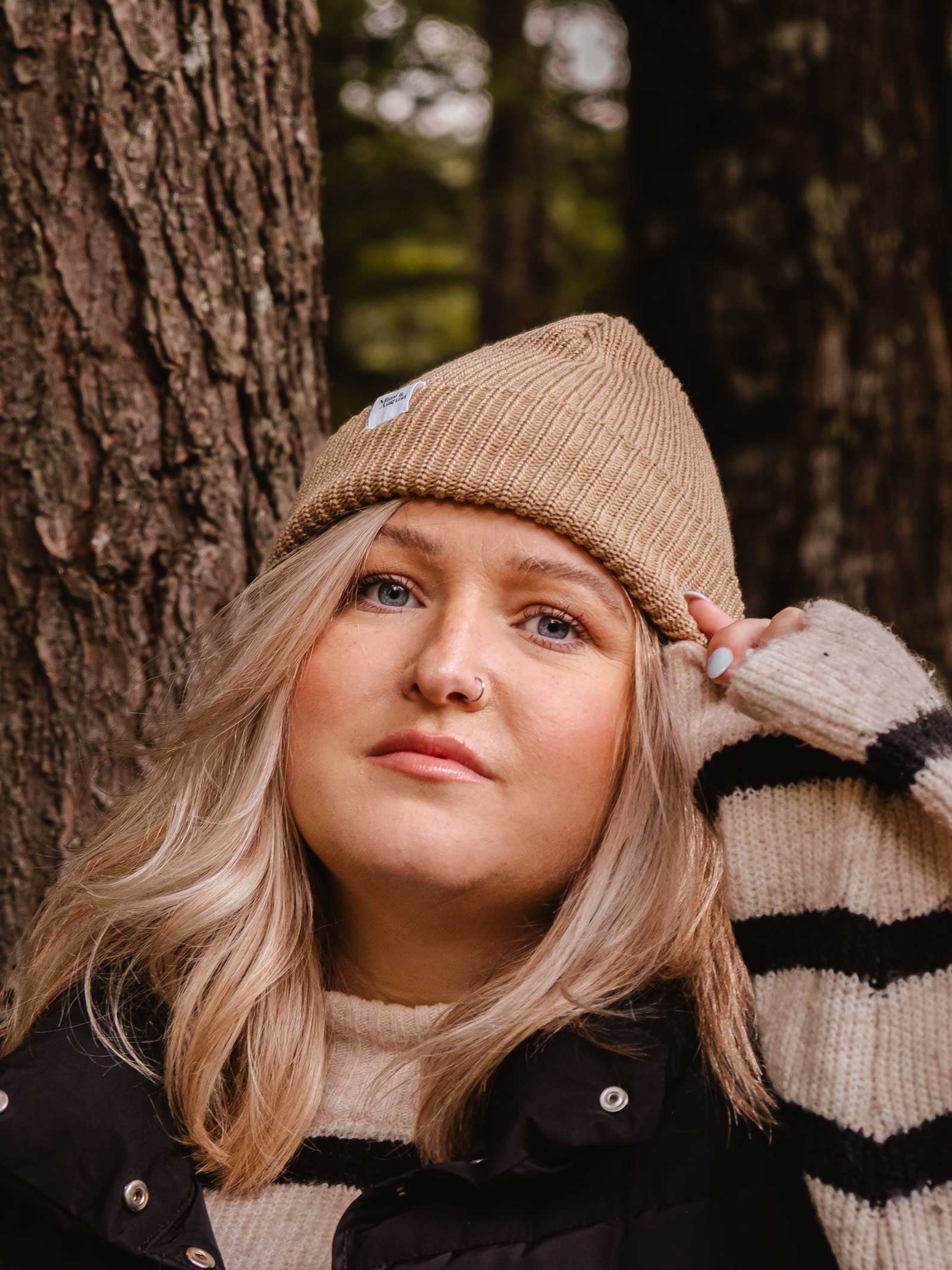 A person is standing next to a tree, wearing a black jacket over a striped sweater and topped with the Khaki Cap Super Puff Beanie by Mimi & August, making for the perfect winter accessory.