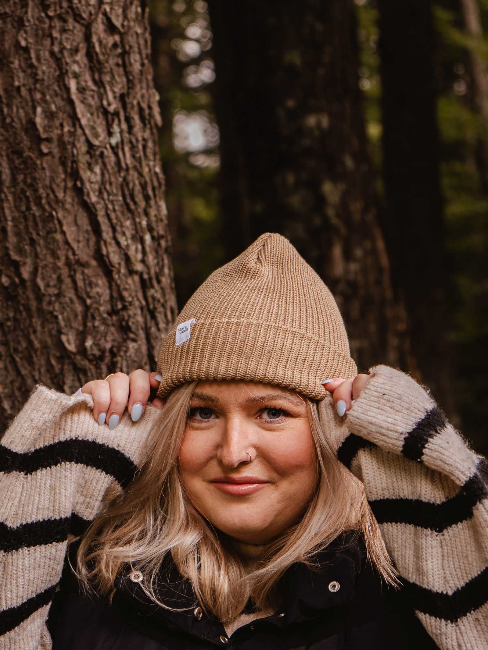 A person wearing a Mimi & August Khaki Cap Super Puff Beanie and a striped sweater stands in front of a tree in a forest, showcasing the perfect winter accessory.