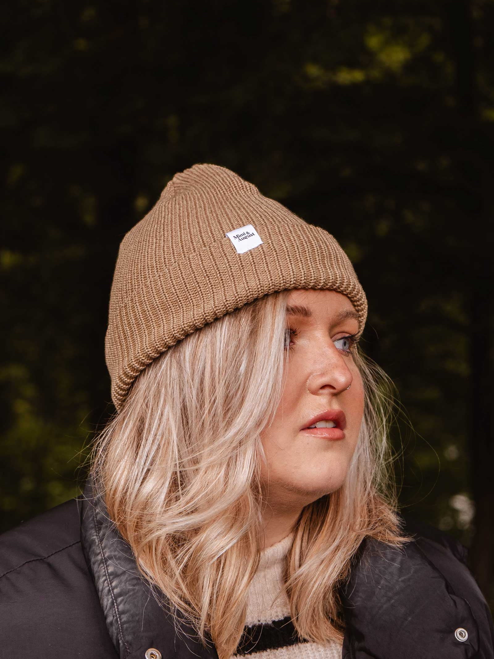 A person with blonde hair wearing a Mimi & August Khaki Cap Super Puff Beanie and a black jacket, perfect as a winter accessory, is glancing to the side against a blurred outdoor background.