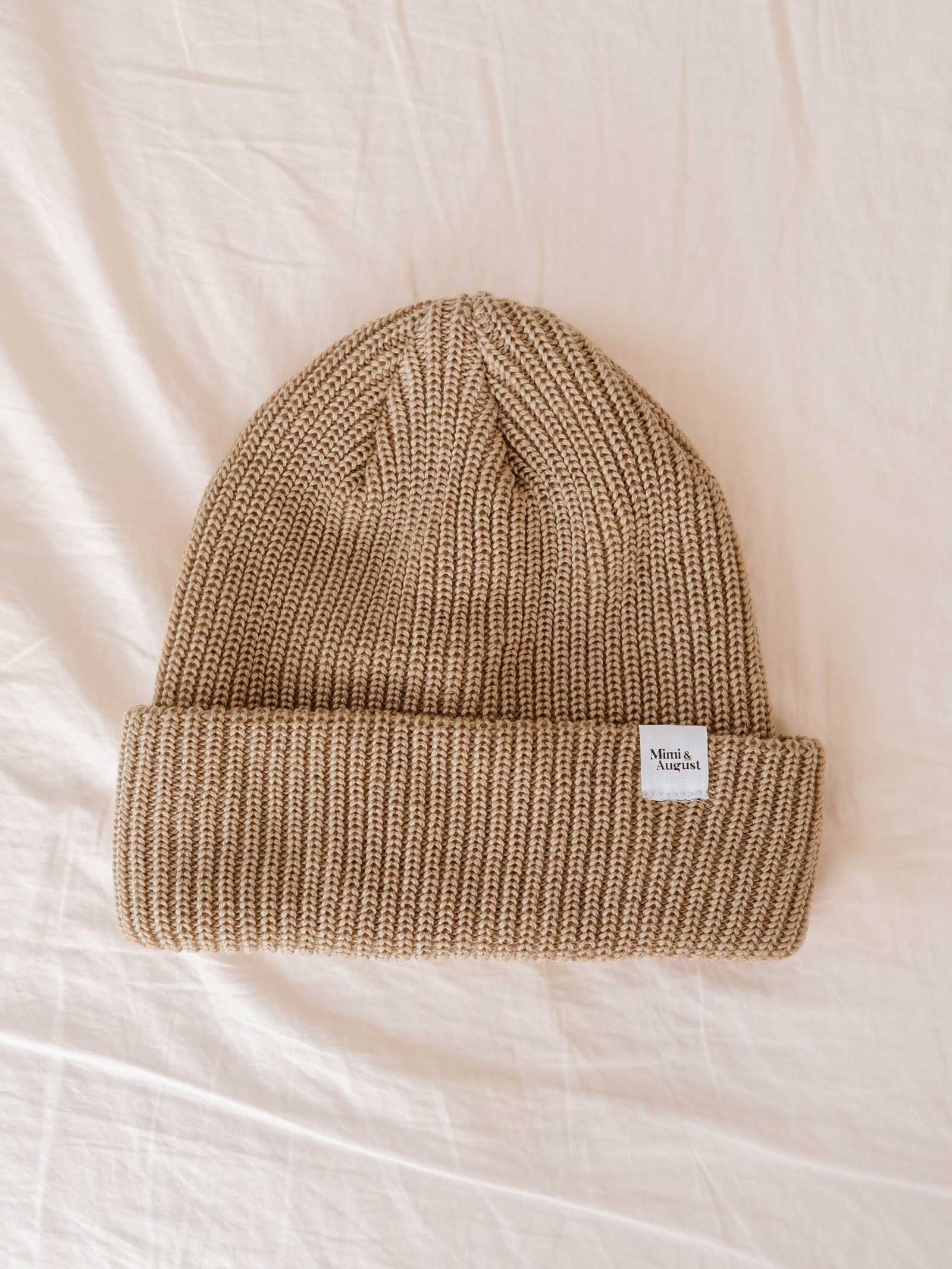 A folded light brown knitted beanie hat, the Khaki Cap Super Puff Beanie by Mimi & August, with a small white tag on the brim, rests on a cream-colored fabric background. This unisex knit winter accessory seamlessly blends style and warmth.