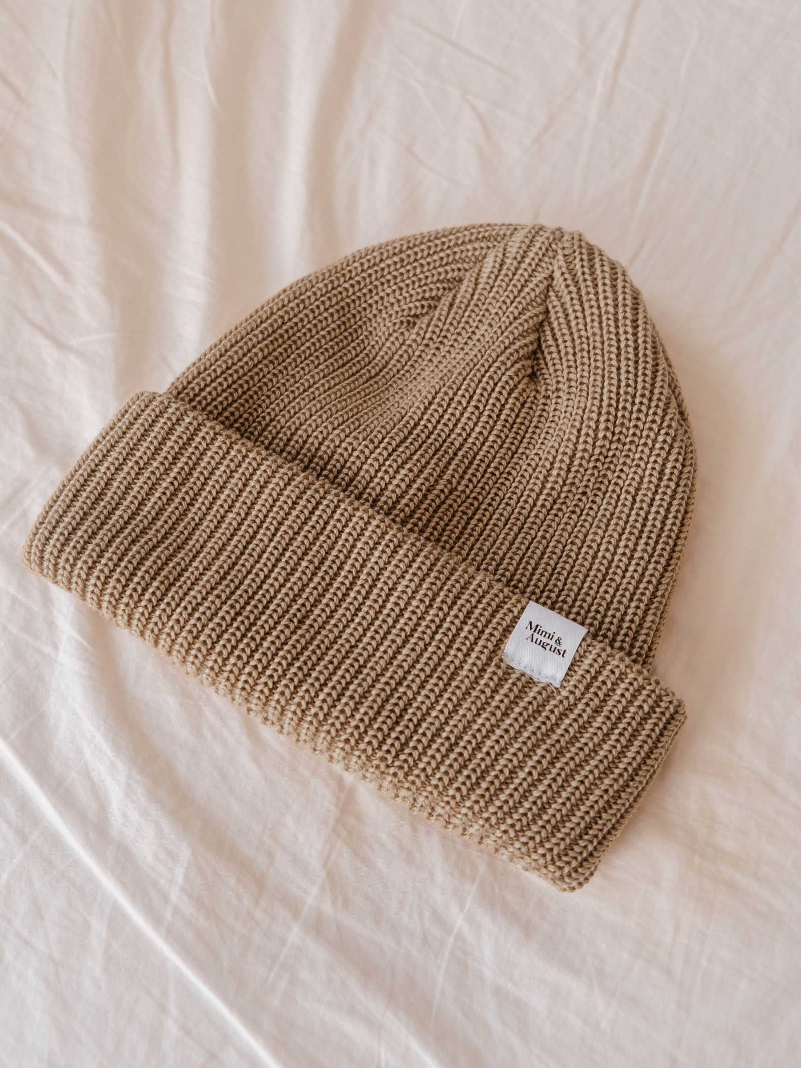 The Khaki Cap Super Puff Beanie, a stylish beige knit winter accessory from Mimi & August, features a small white tag displaying text and lies on a light-colored fabric surface. This unisex piece is perfect for staying warm in style.