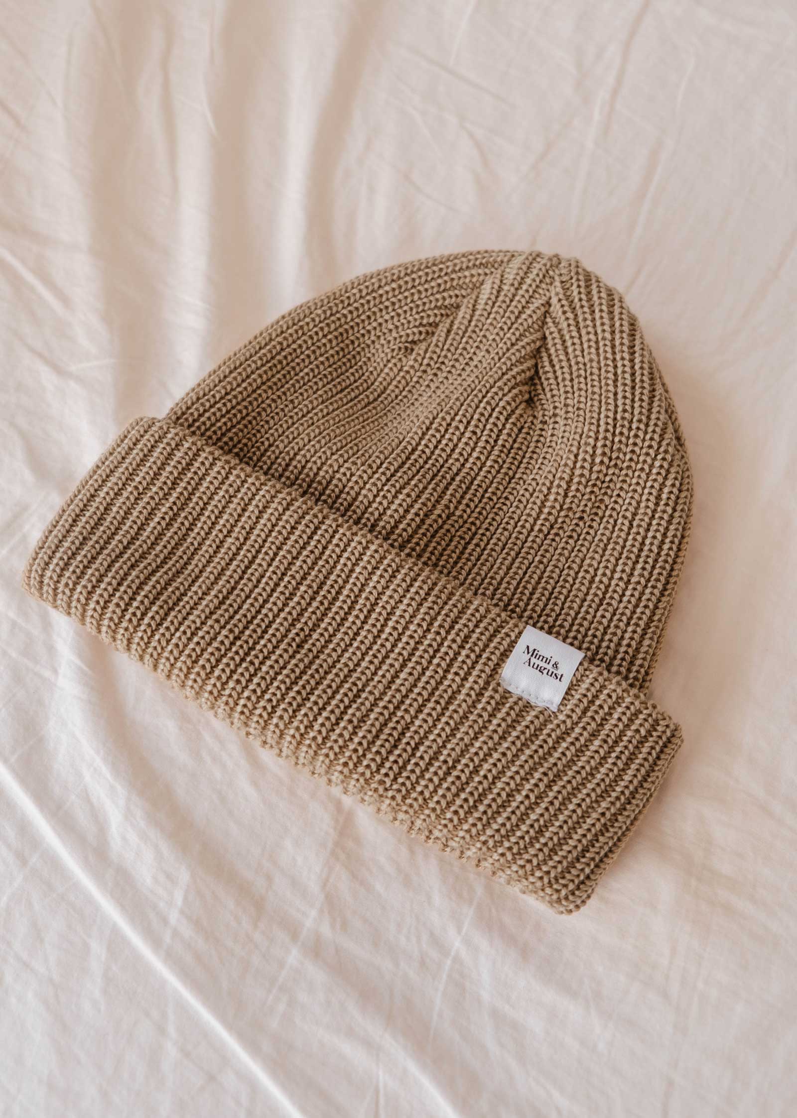 The Khaki Cap Super Puff Beanie, a stylish beige knit winter accessory from Mimi & August, features a small white tag displaying text and lies on a light-colored fabric surface. This unisex piece is perfect for staying warm in style.