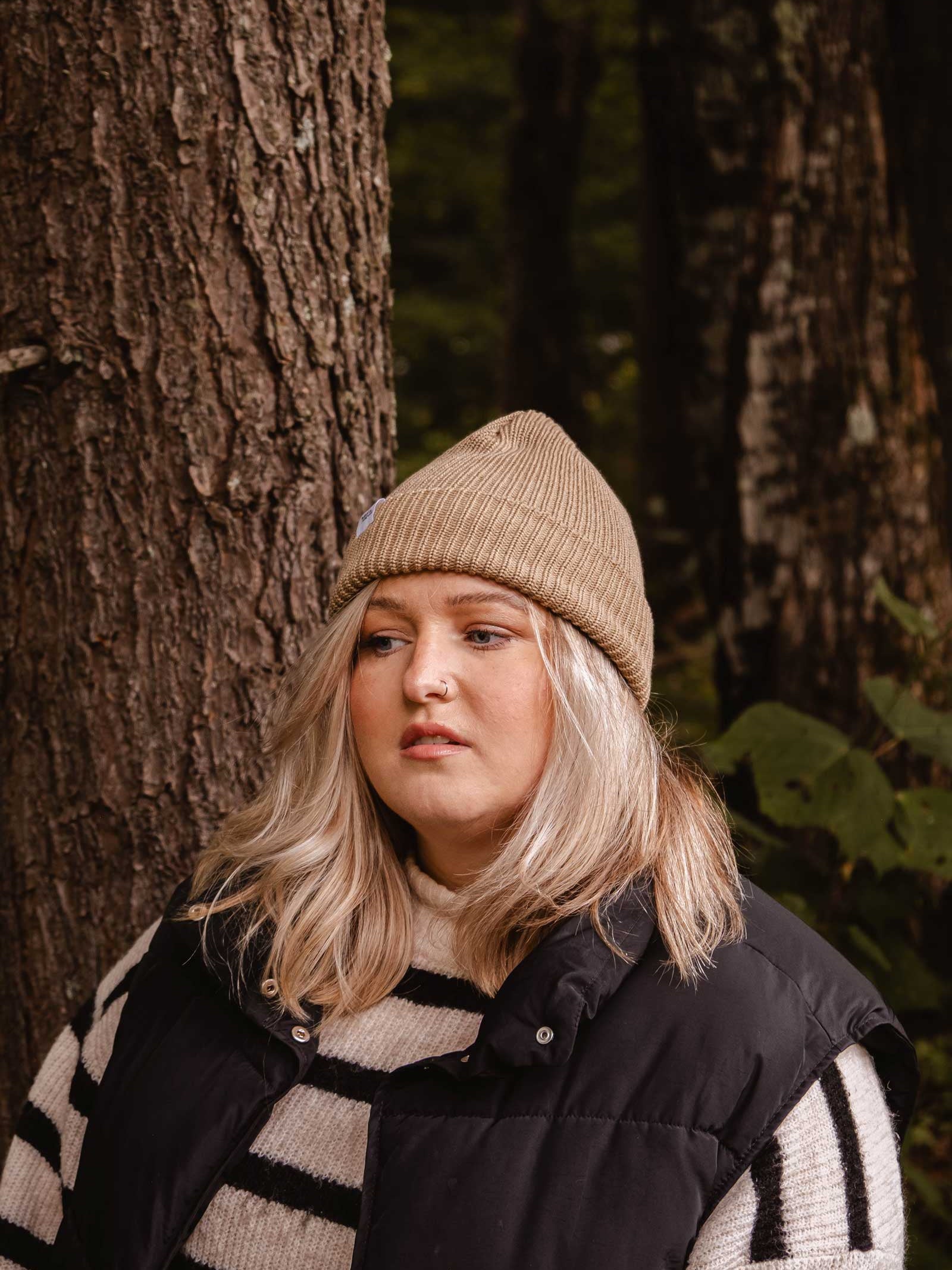 A person with long, blonde hair wearing a Mimi & August Khaki Cap Super Puff Beanie, a black puffer vest, and a striped sweater stands in front of a tree in a forest setting.