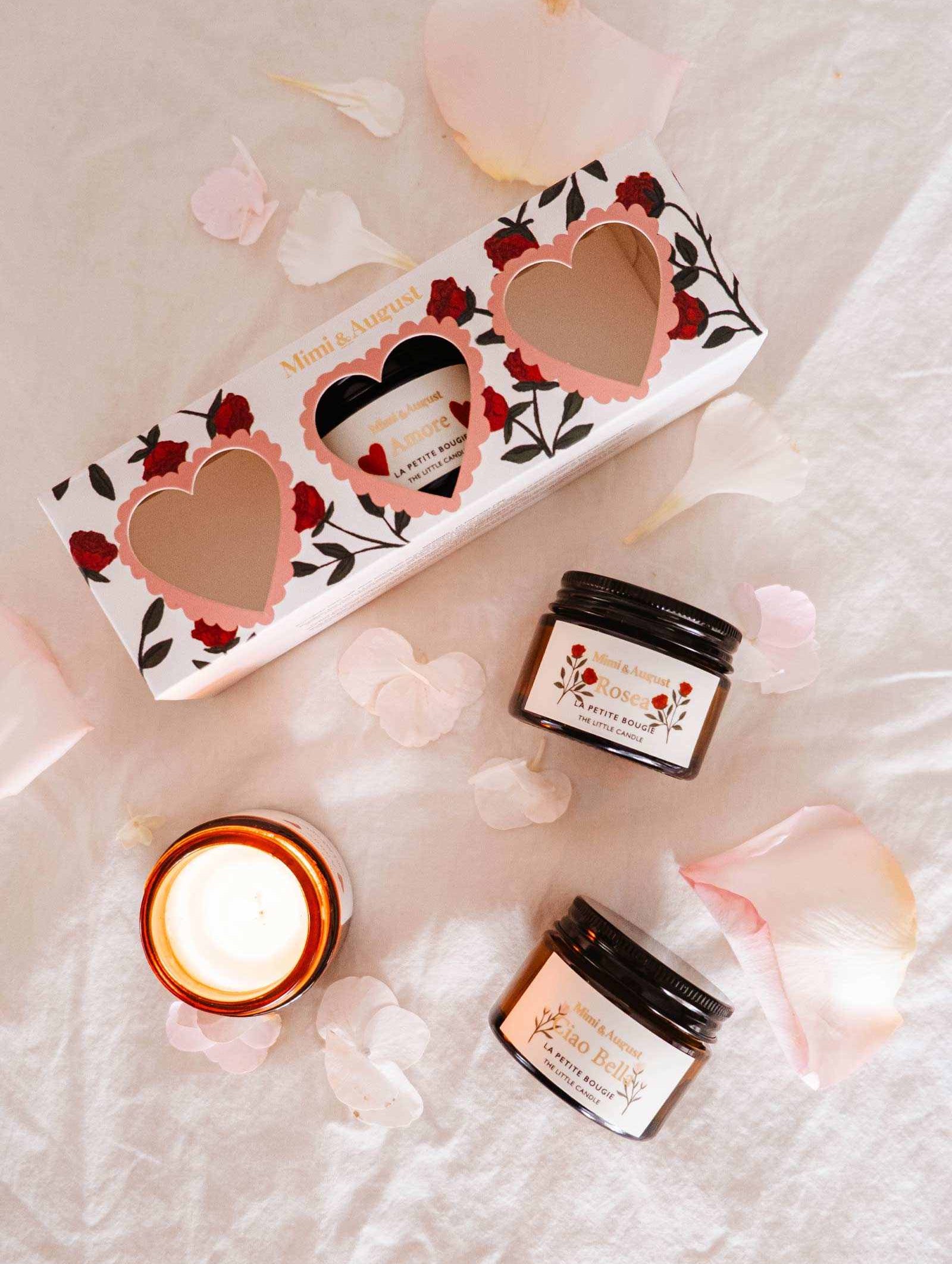 Skincare items, including two jars and an elegant candle, rest on soft pink fabric with scattered petals next to the Mimi & August La Romance Box Set featuring heart-shaped cutouts. With exclusive fragrances, this collection adds luxury to your beauty routine.
