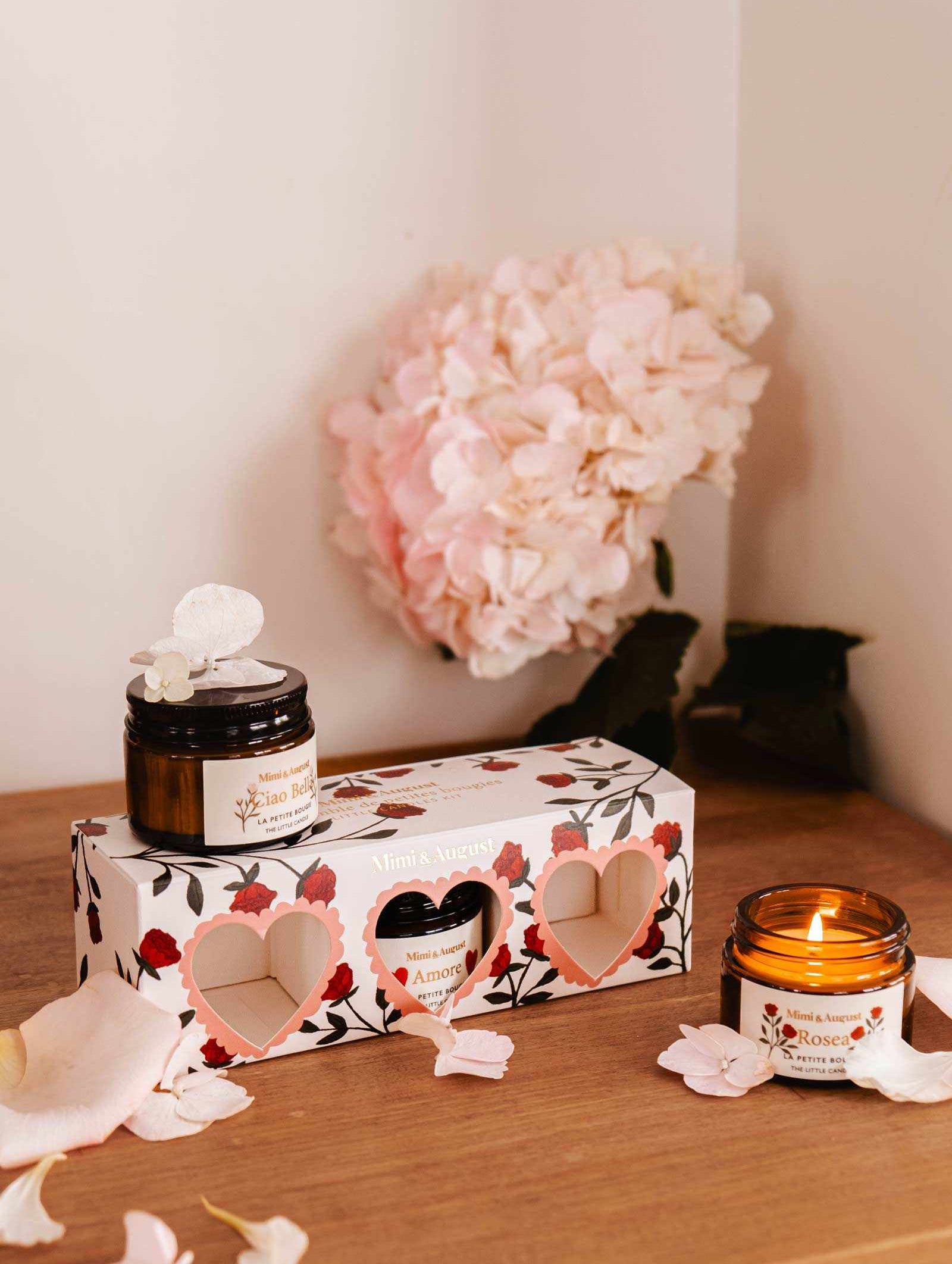 Two elegant candles in glass jars, one lit, adorn a wooden surface beside the Mimi & August La Romance Box Set. Exclusive fragrances fill the air as pink hydrangeas and scattered petals add floral charm to the scene.